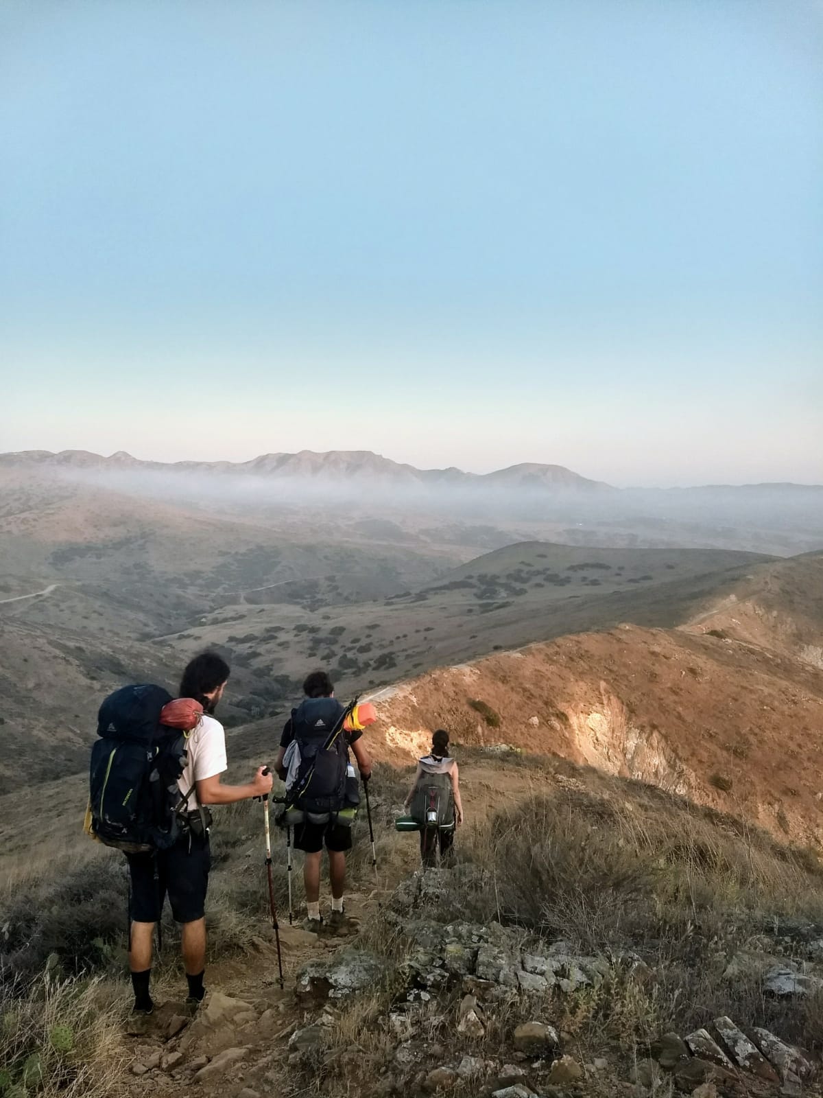 Three people with backpacks on hike down a mountain outside.