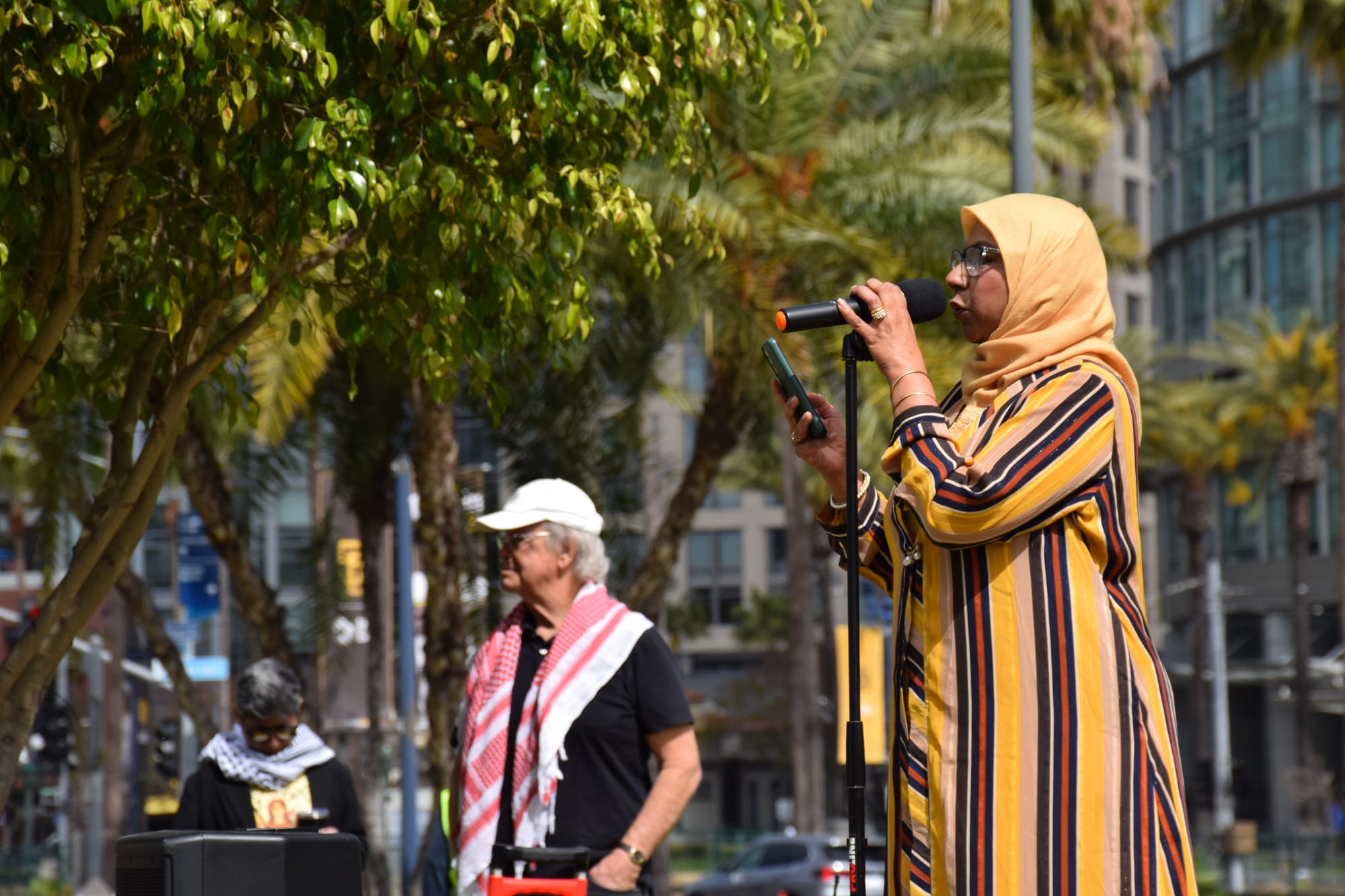 A woman in a yellow hijab speaks into a microphone outside in front of a tree and two other people