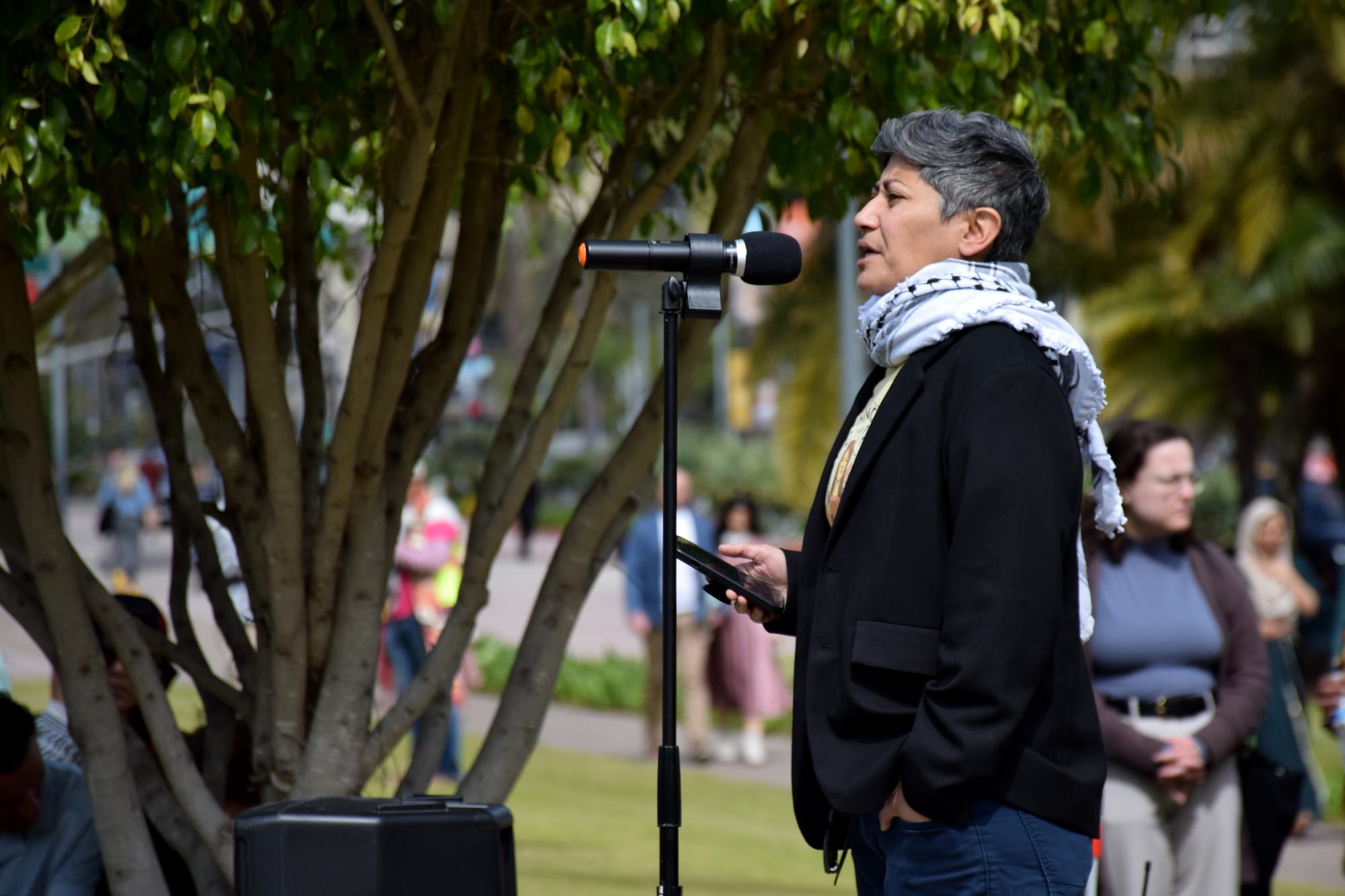 A woman with short hair wearing a keffiyeh speaks at a microphone in a park with people walking on the path behind her.