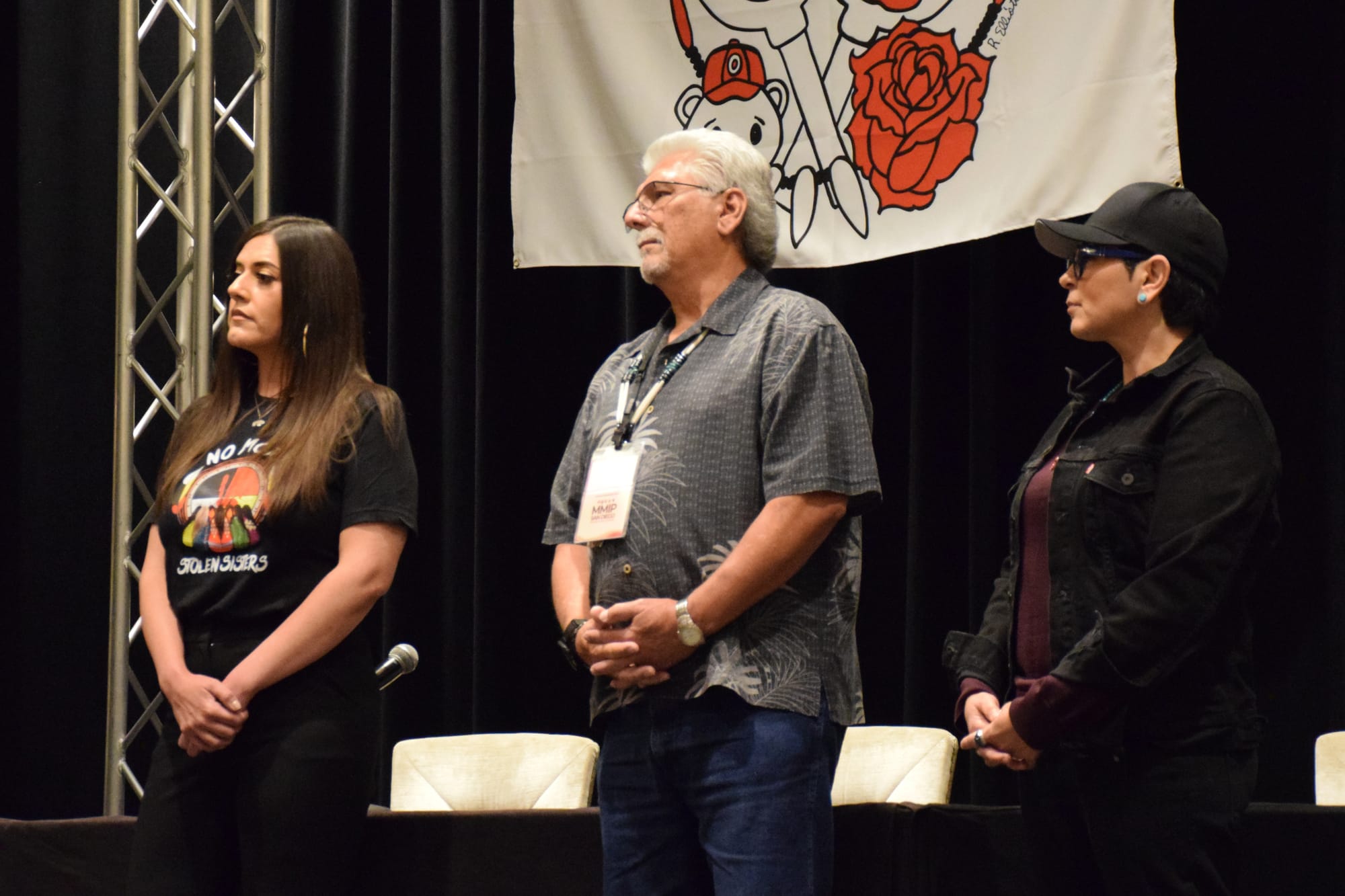 Two women and a man stand on a stage looking at someone speaking who is not in the image