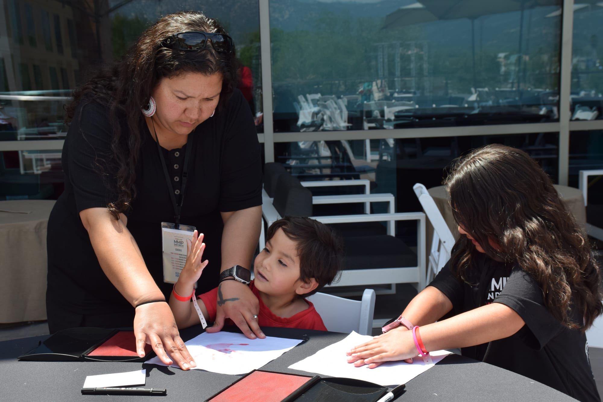 A woman helps her two kids with a craft project sitting outside at a large table