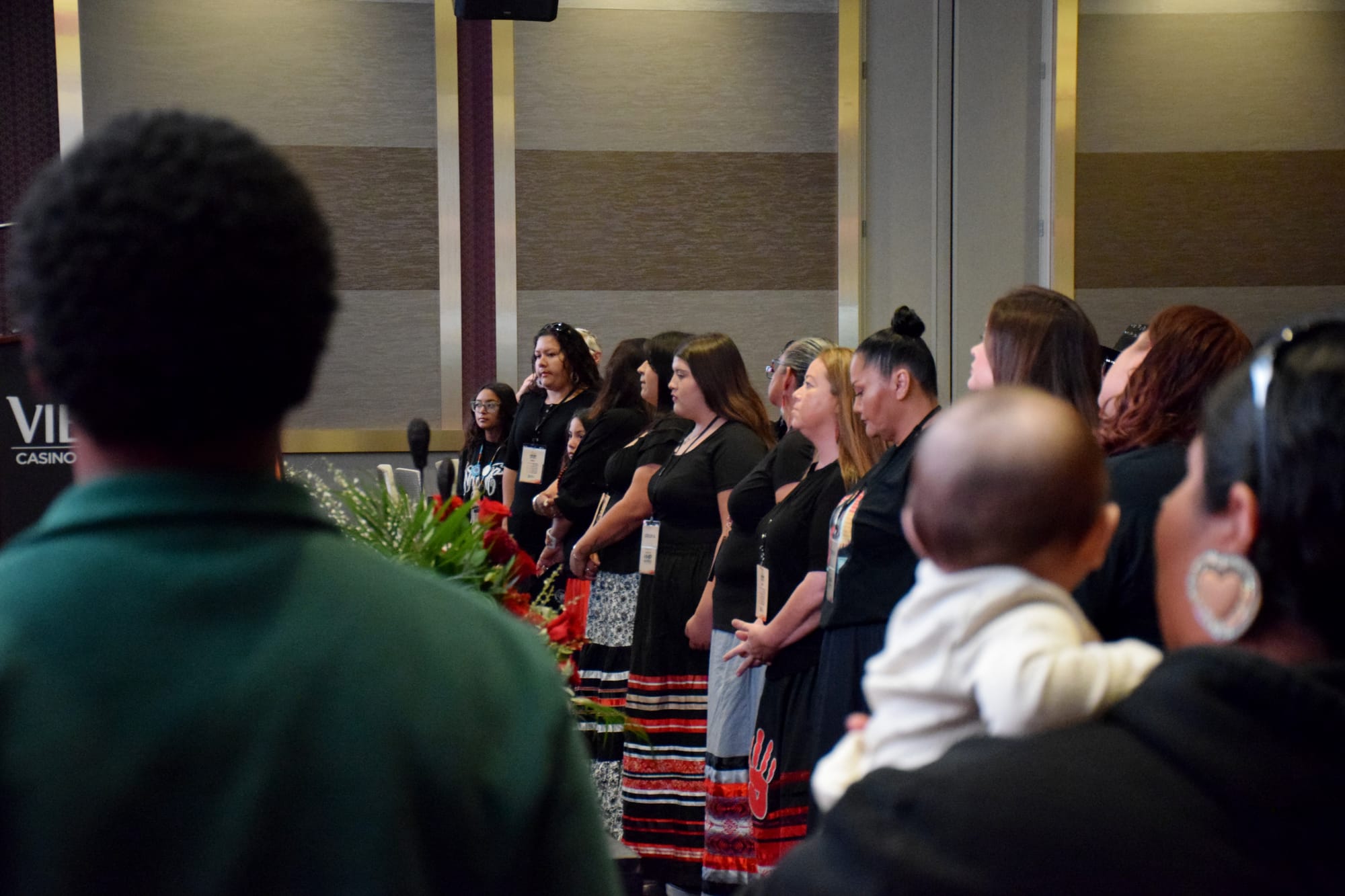 A line of people wearing black t-shirts and red and black ribbon skirts stand in a conference room