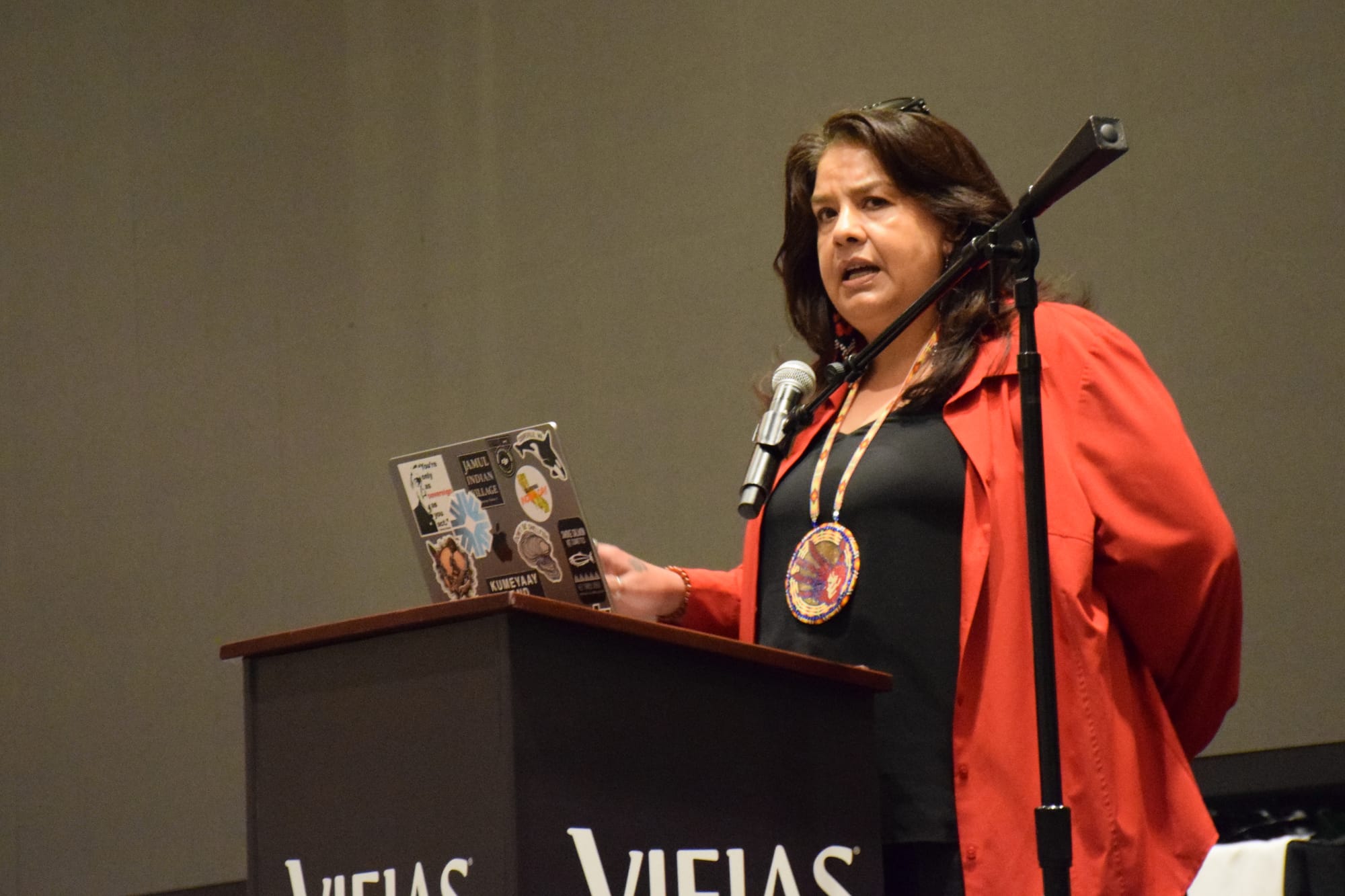 A woman wearing a red button up over a black t-shirt stands on a stage behind a podium speaking