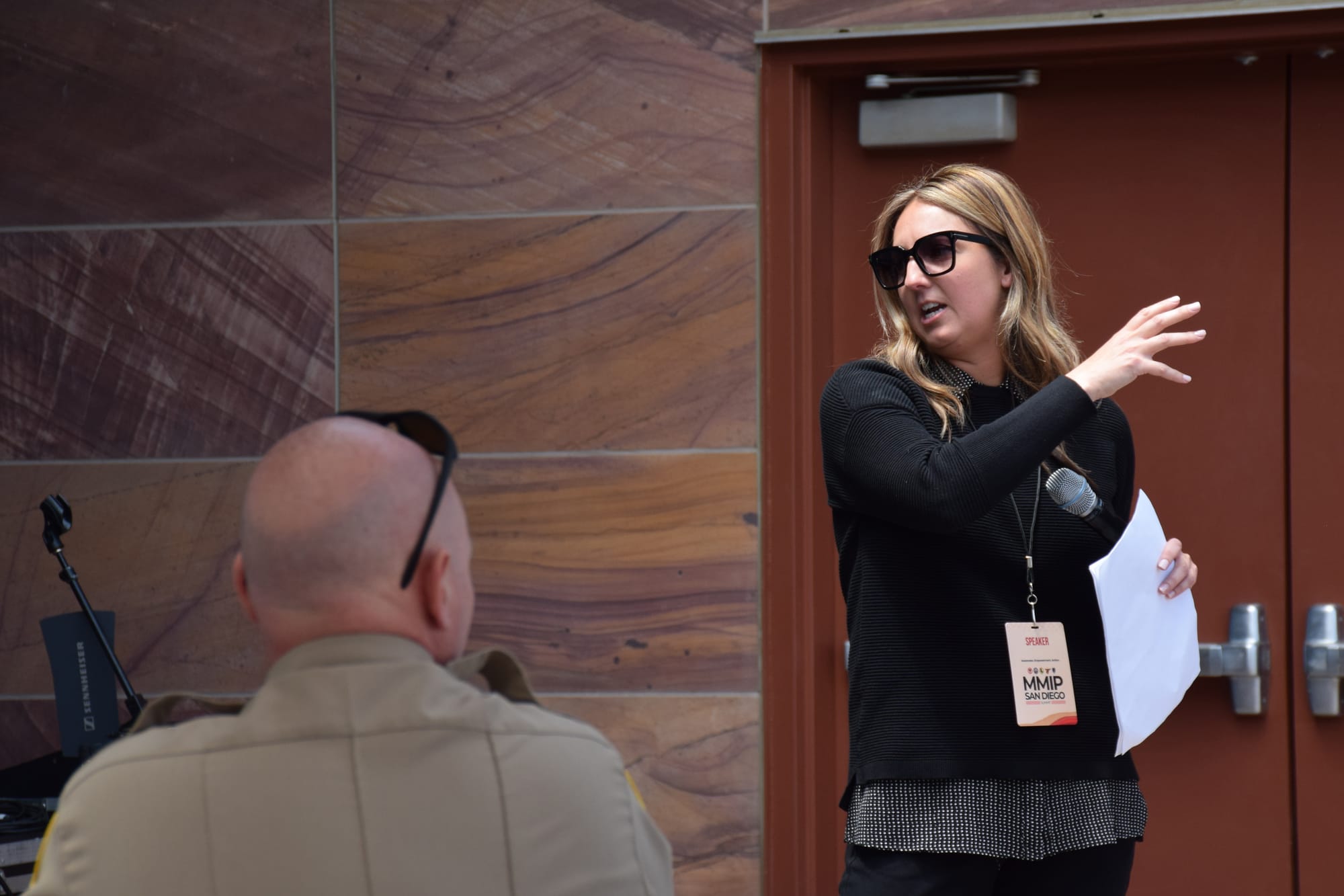 A woman wearing sunglasses holding a microphone talks outside in front of a sheriff's deputy