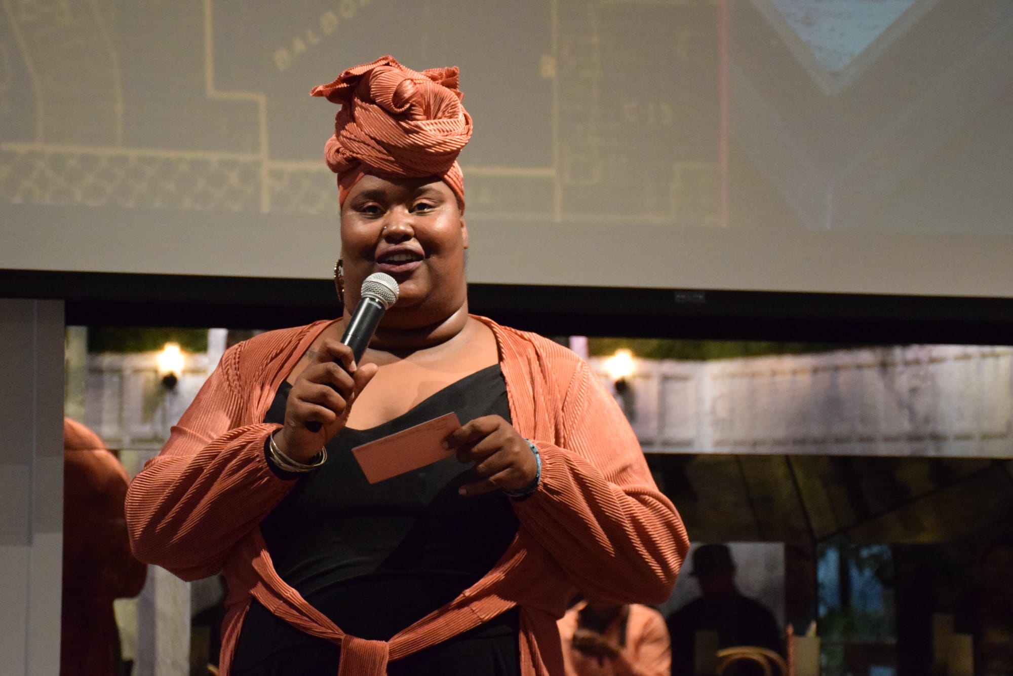 A woman with an orange-red headwrap and jacket and black top speaks into a microphone