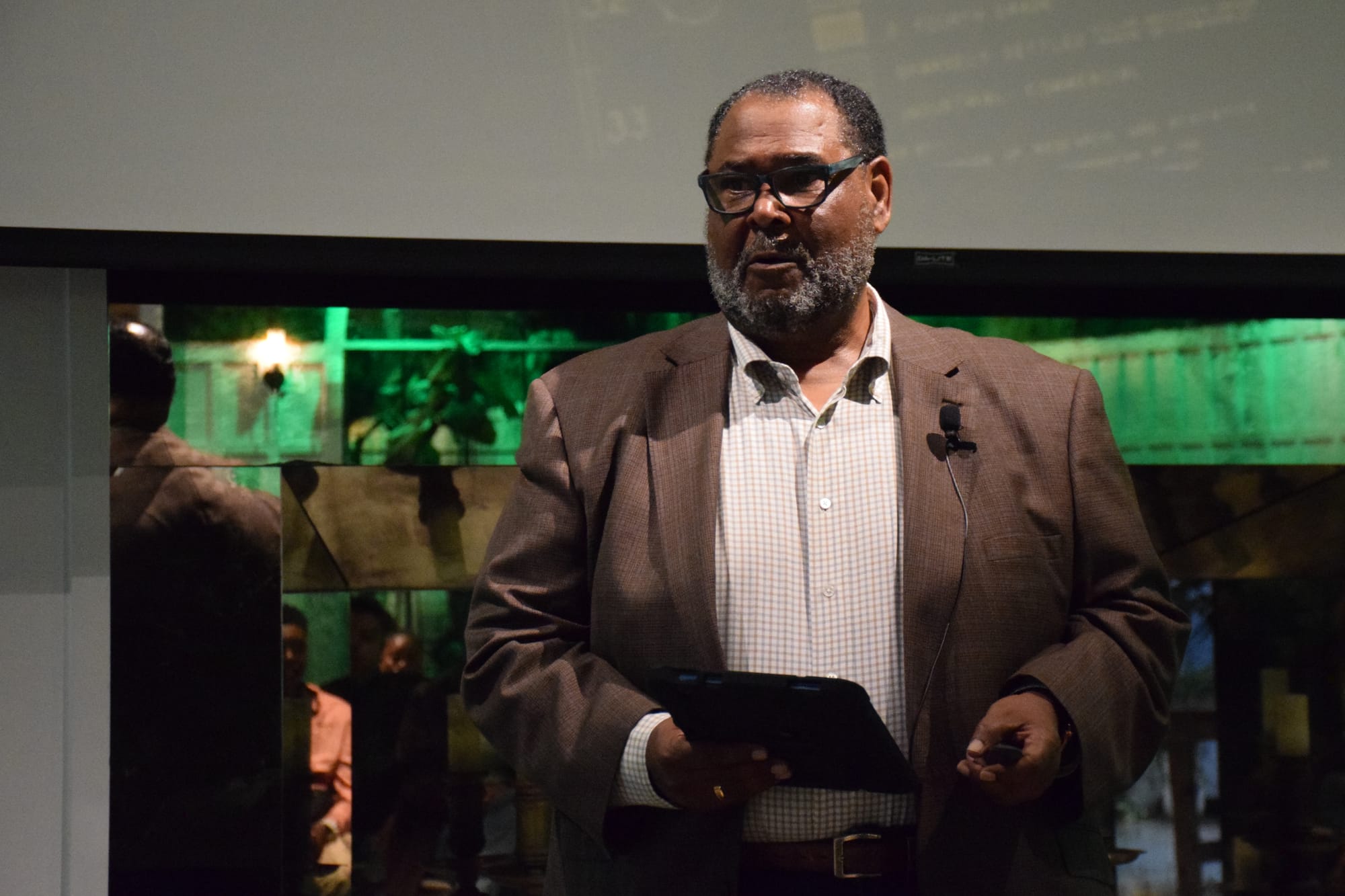 A man wearing glasses and a brown suit jacket speaks with a green light reflecting behind him