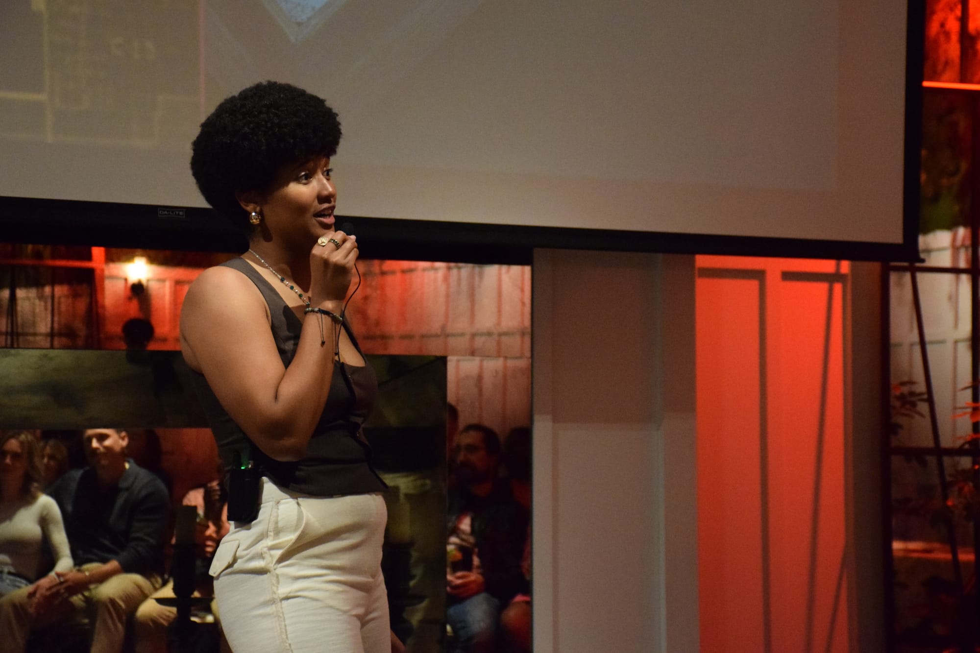 A woman in a brown top and cream pants holds a lavaliere microphone as she speaks, and red light reflects off the wall behind her
