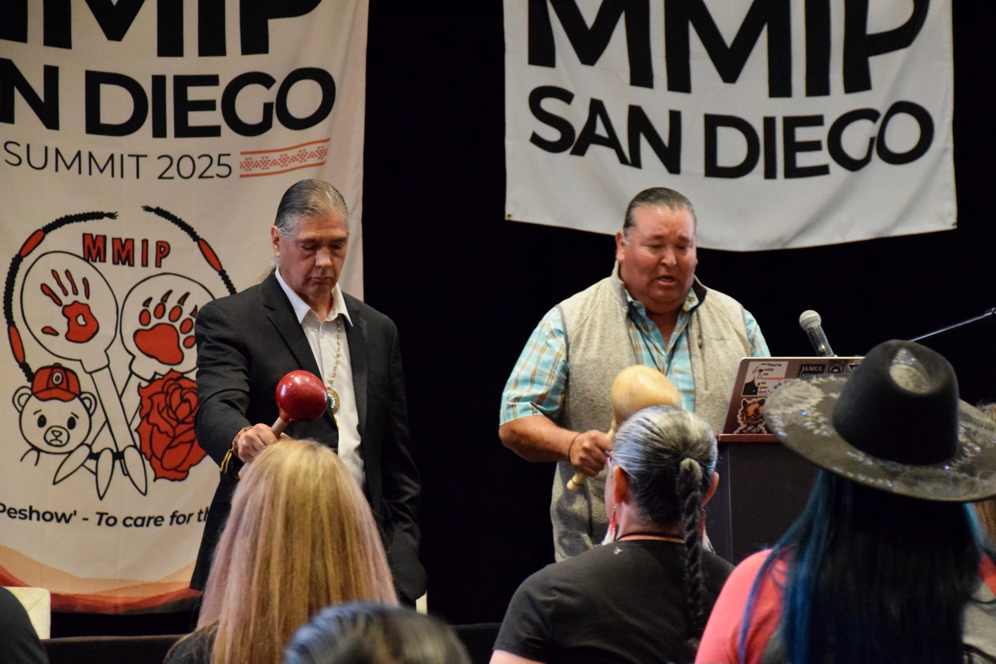 Two men stand on a stage in front of a crowd of people holding gourd rattles and singing with two signs that say MMIP San Diego behind them.