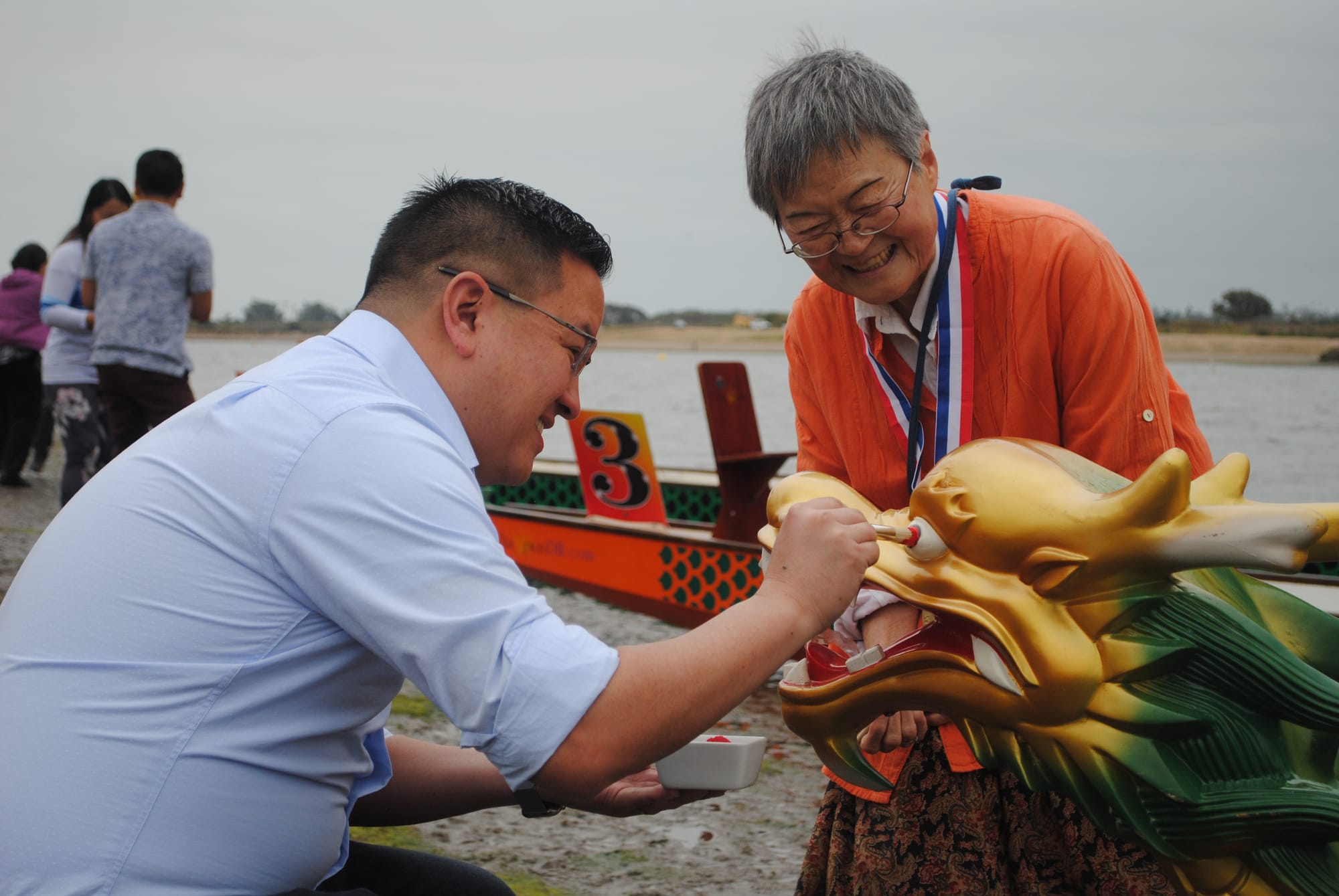 A man wearing a light blue shirt kneels and holds a paint brush to a dragon’s eye while a woman wearing an orange blouse leans over and watches smiling