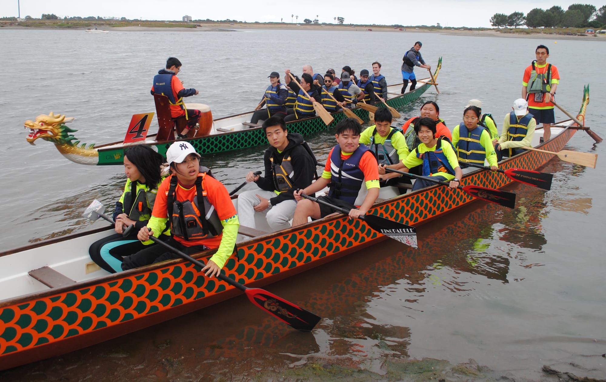 A long slender boat with orange and green painted scales carries eleven children who are holding paddles and wearing neon yellow jerseys and life vests