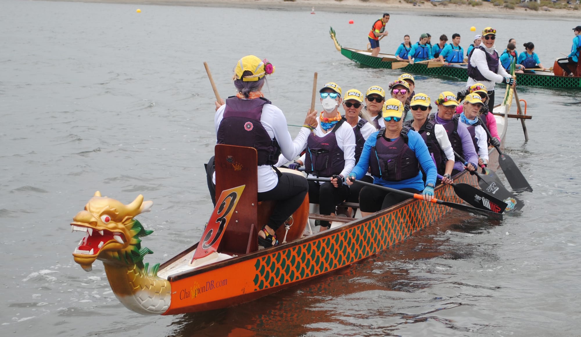 A long slender boat with orange and green painted scales carries eleven women who are holding paddles and wearing yellow hats and life vests