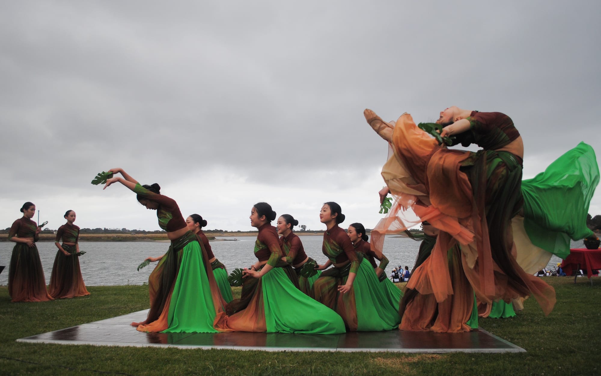 A dance group of young women wearing flowing, brown, green and orange dance attire hold a pose upon a stage. One woman in the foreground leaps while arching her back