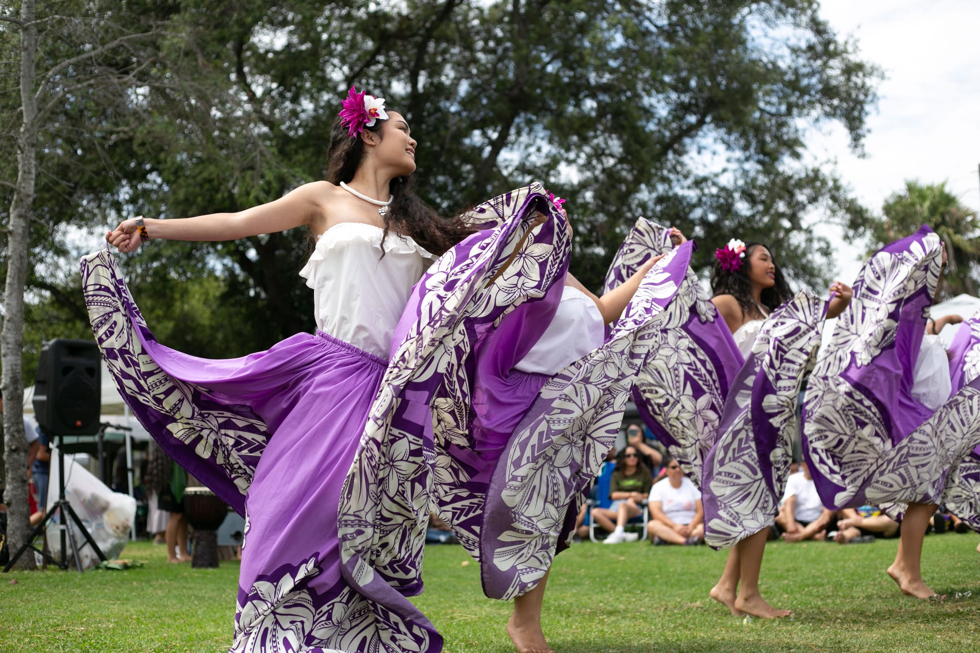 Four women wearing purple, patterned skirts, white shirts and flowers in their hair dance in front of a tree on the grass at an event with people sitting in the background