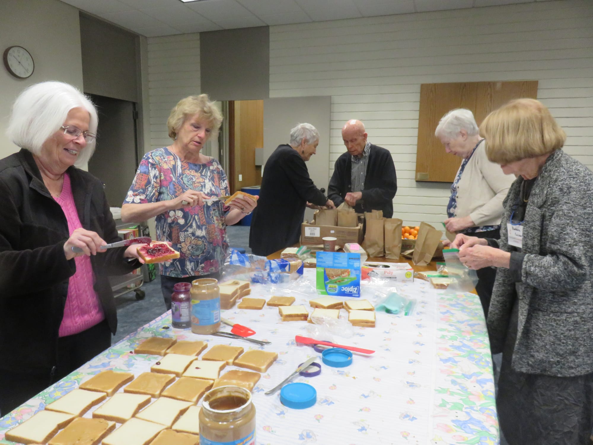 Two women make peanut butter and jelly sandwiches on the left side of a large table while four other people bag the sandwiches and place them in paper bags