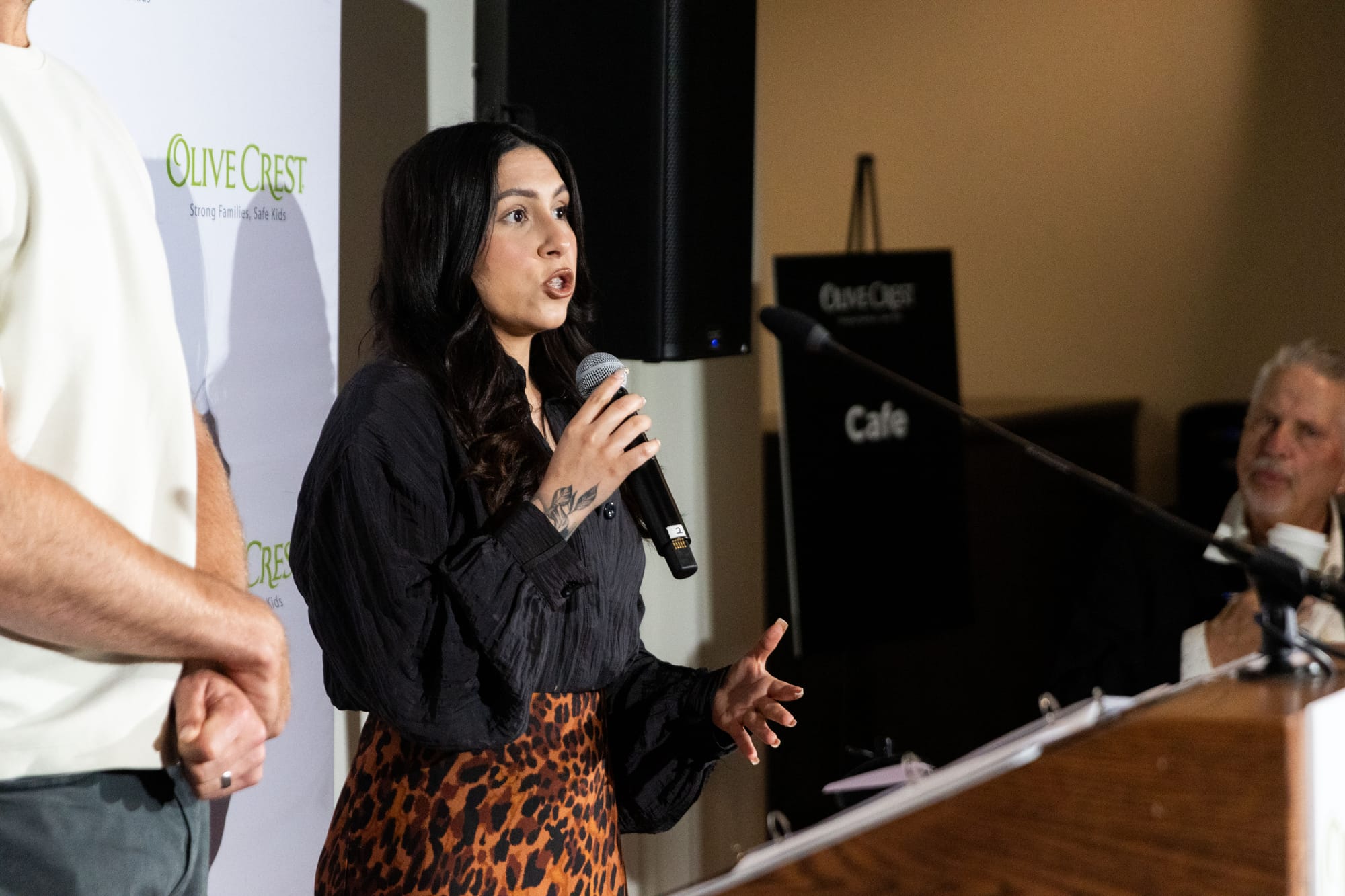 A woman in a black top and brown, animal-print skirt holds a microphone and gestures with her hand as she speaks