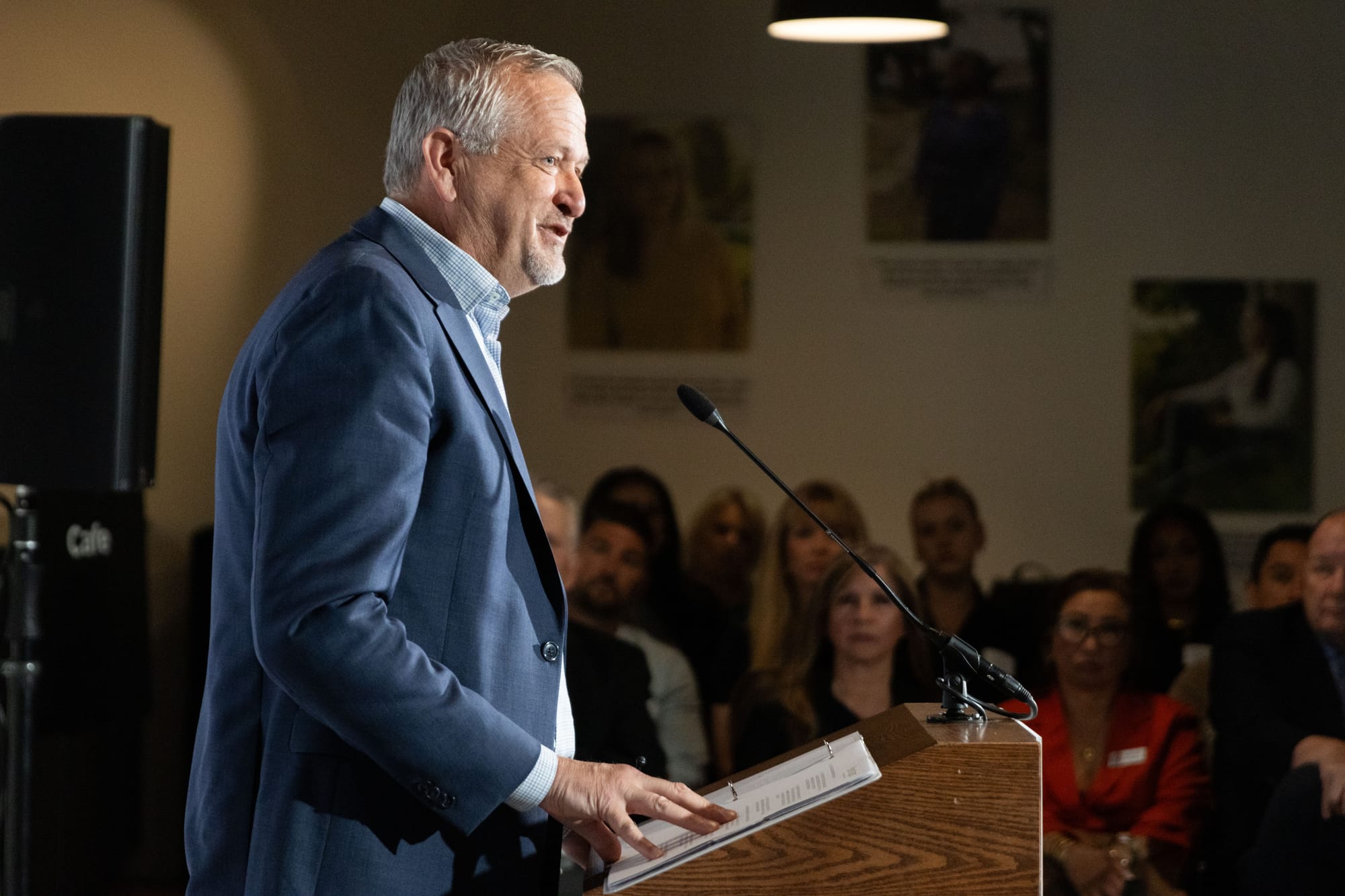 A man in a blue suit seen from the side stands in front of a podium with notes on it, and several faces are out of focus behind him