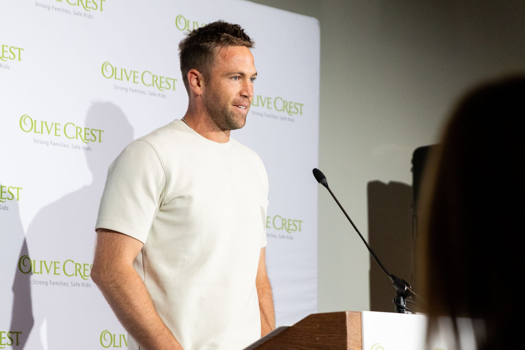 A man in a white shirt stands in front of his podium with a sign that says Olive Crest behind him