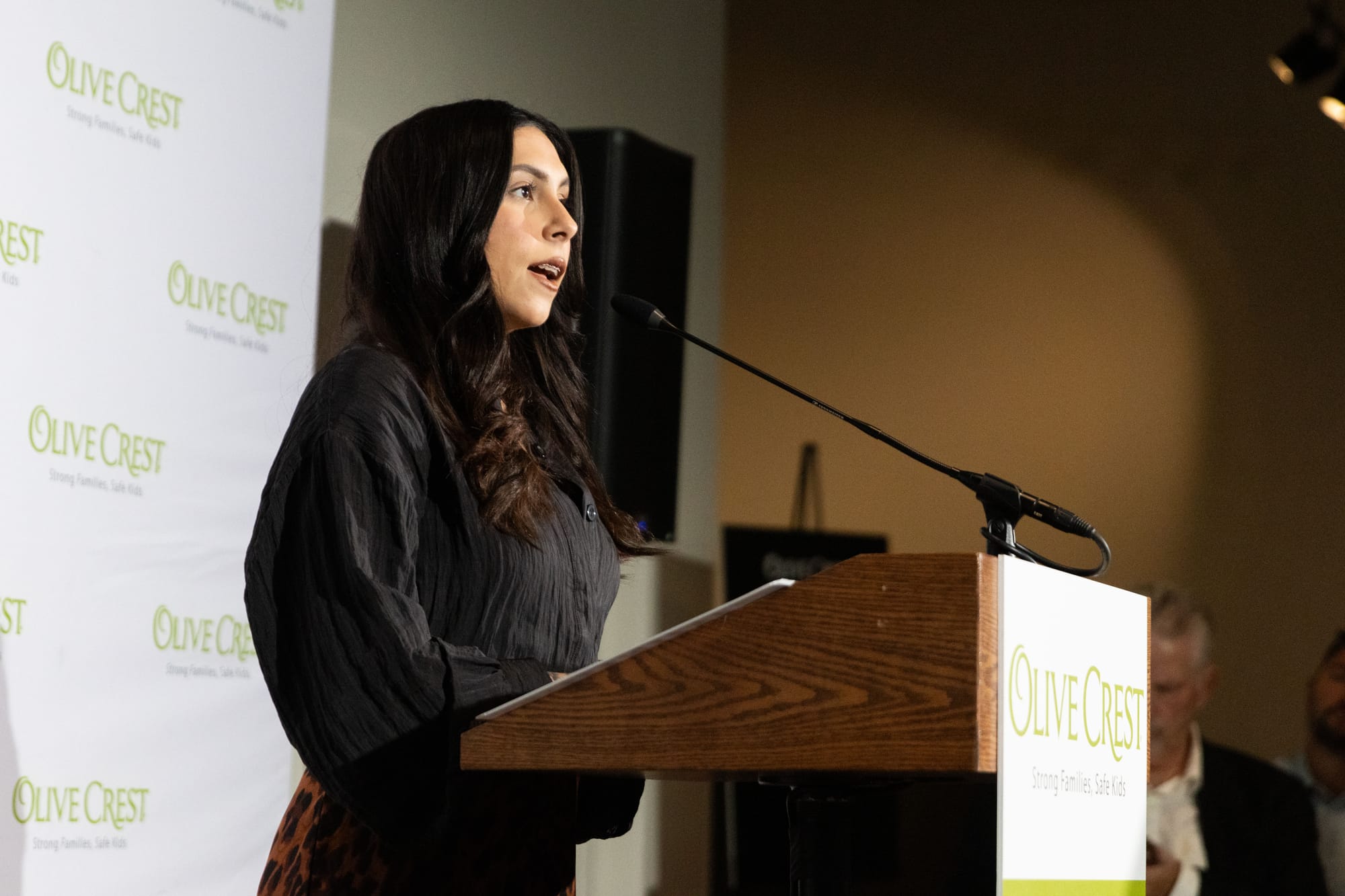 A woman in a black top stands in front of a podium with the Olive Crest logo on it