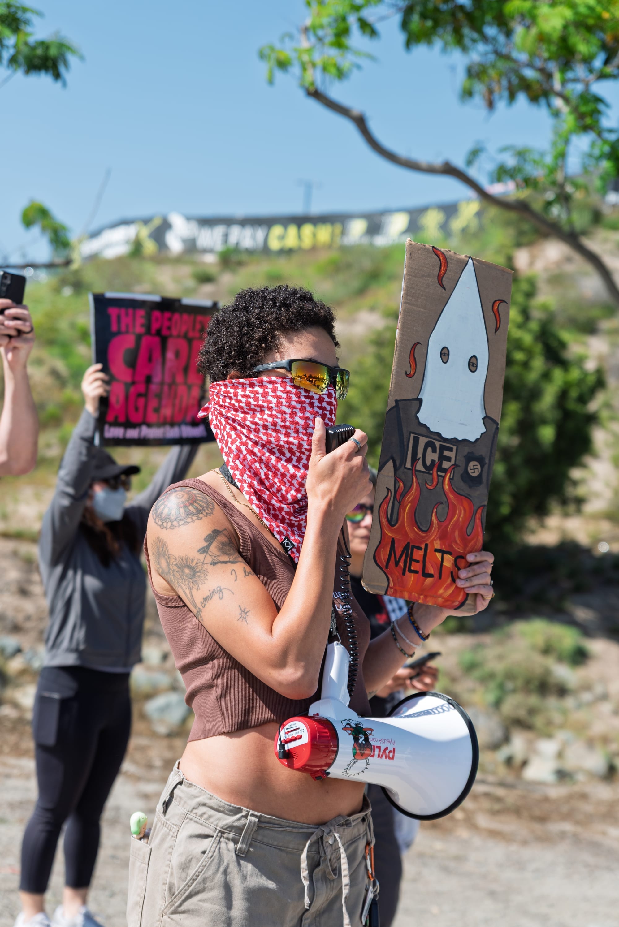 A woman with a brown shirt holds a sign that says, "ICE melts" while speaking into a megaphone