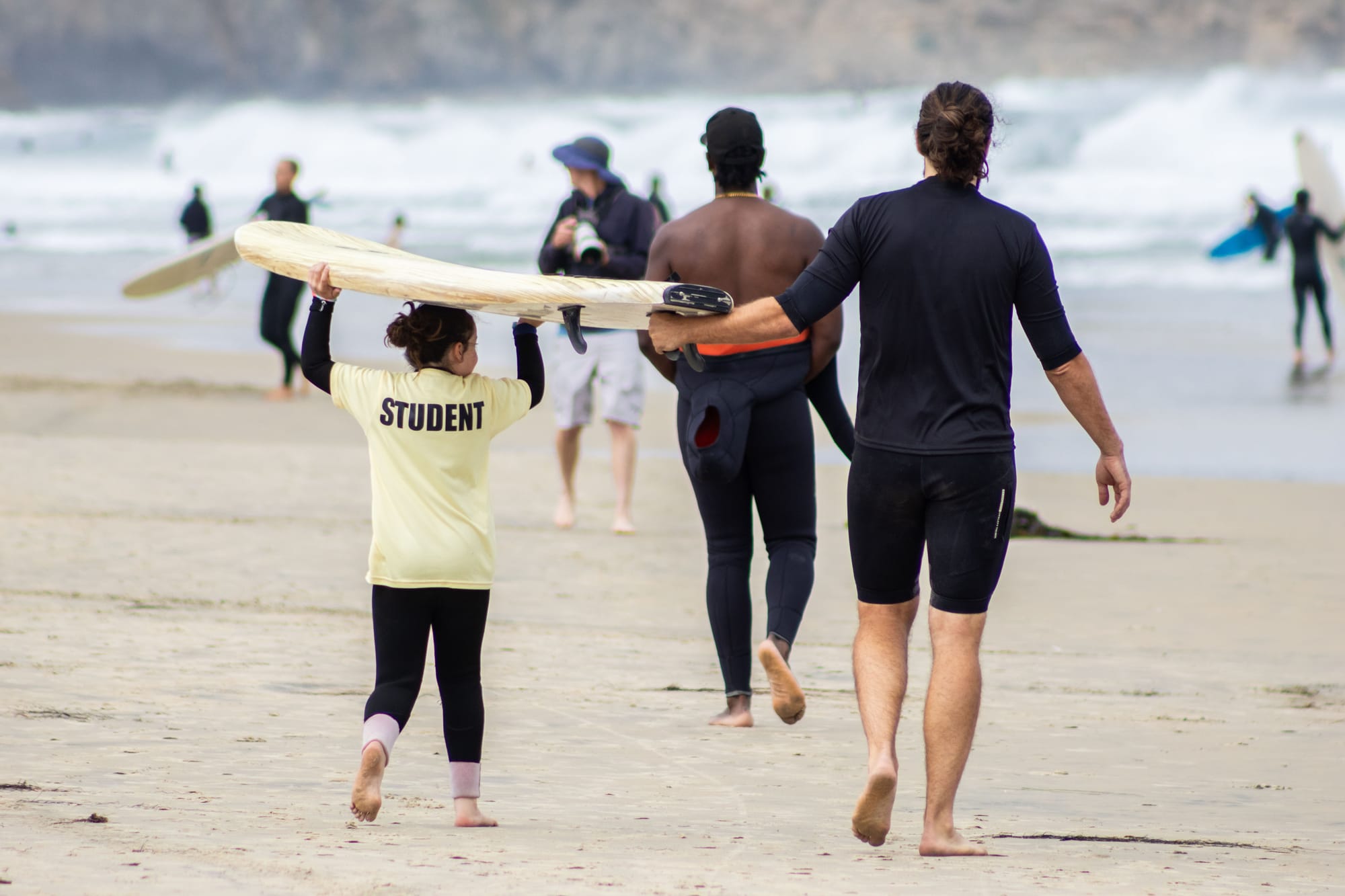 A child holds a surfboard on their head while walking on the beach next to surf instructors