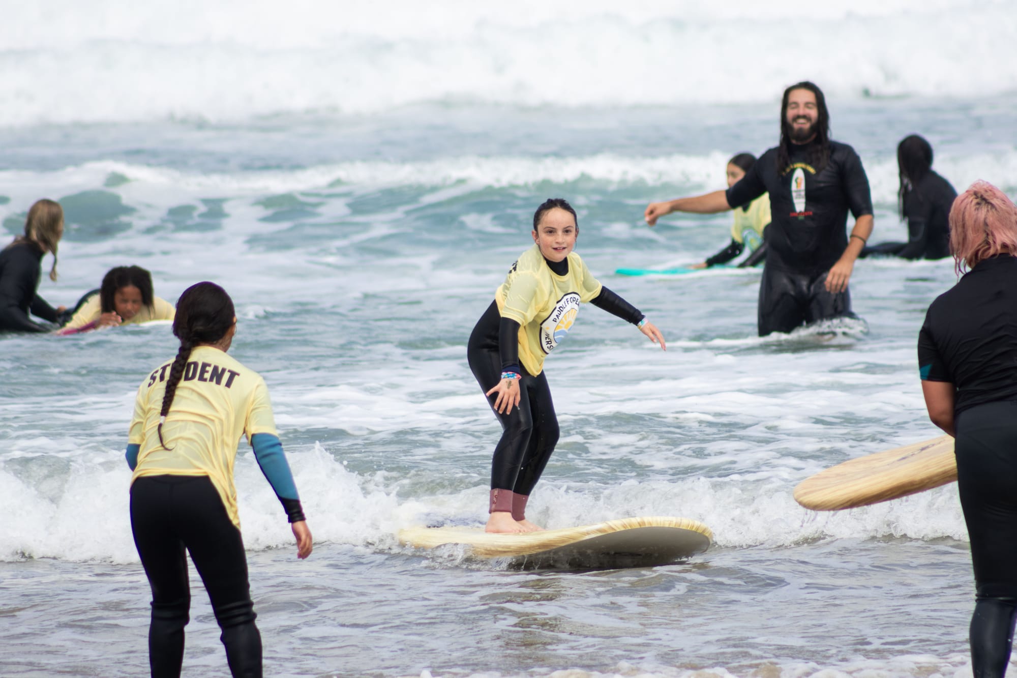Kids practice surfing in the shallow water with instructors around them