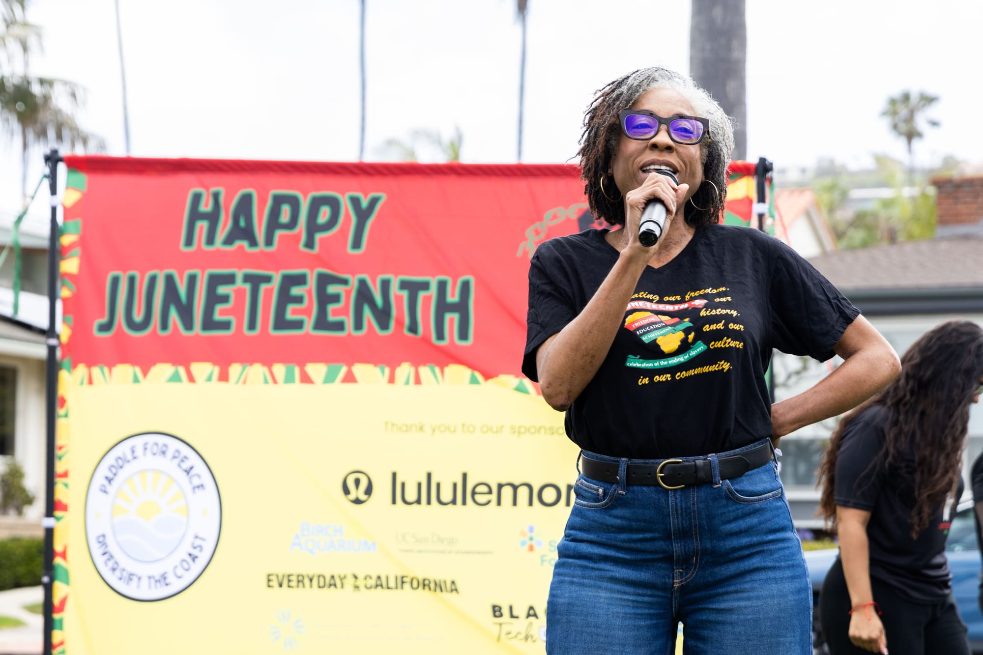 A woman speaks into a microphone outside in front of a sign that reads "Happy Juneteenth"
