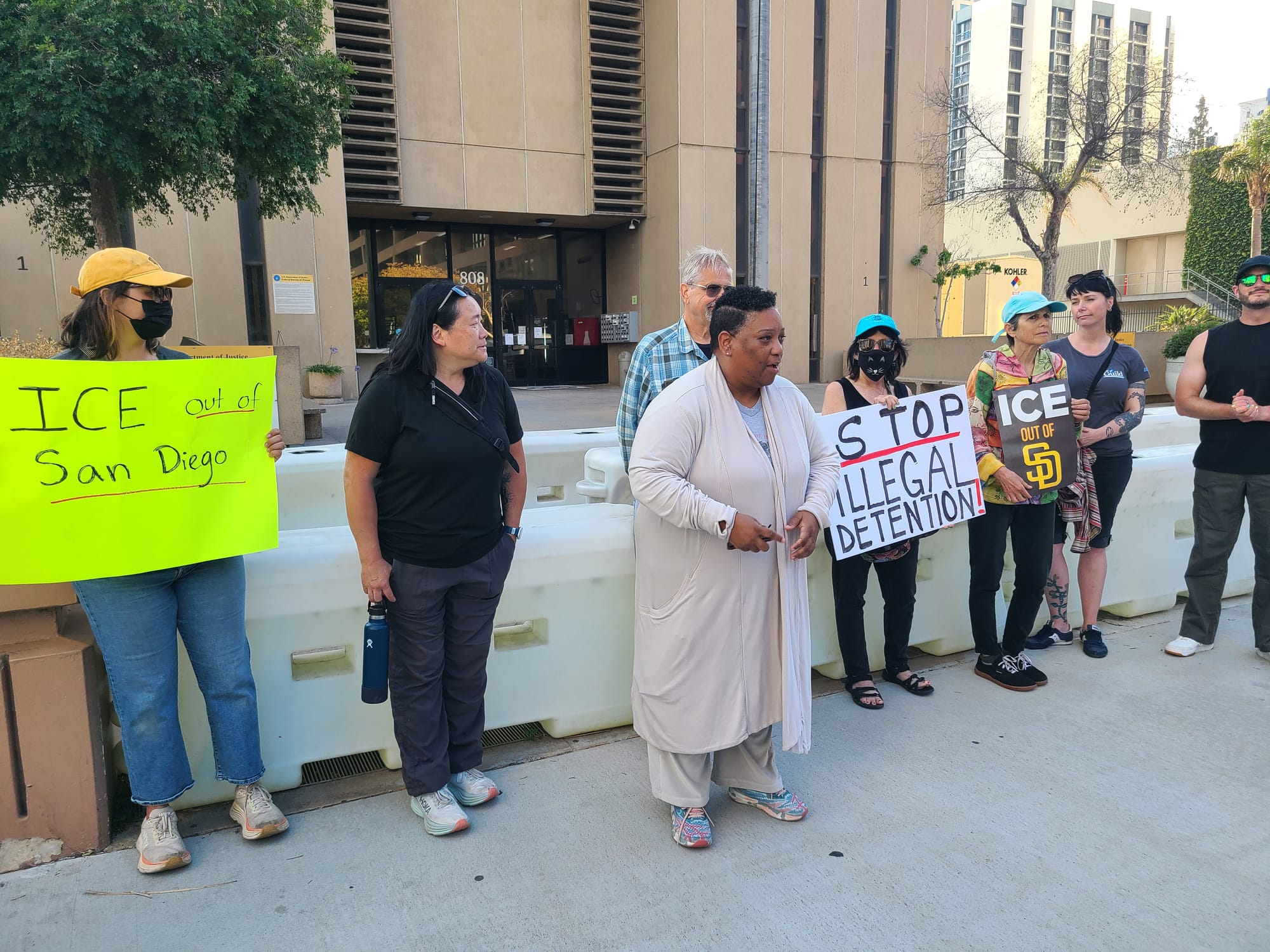 A line up of people stand outside as one person talks in front of a building. Some hold signs reading "ICE out of San Diego" and "Stop illegal detention"