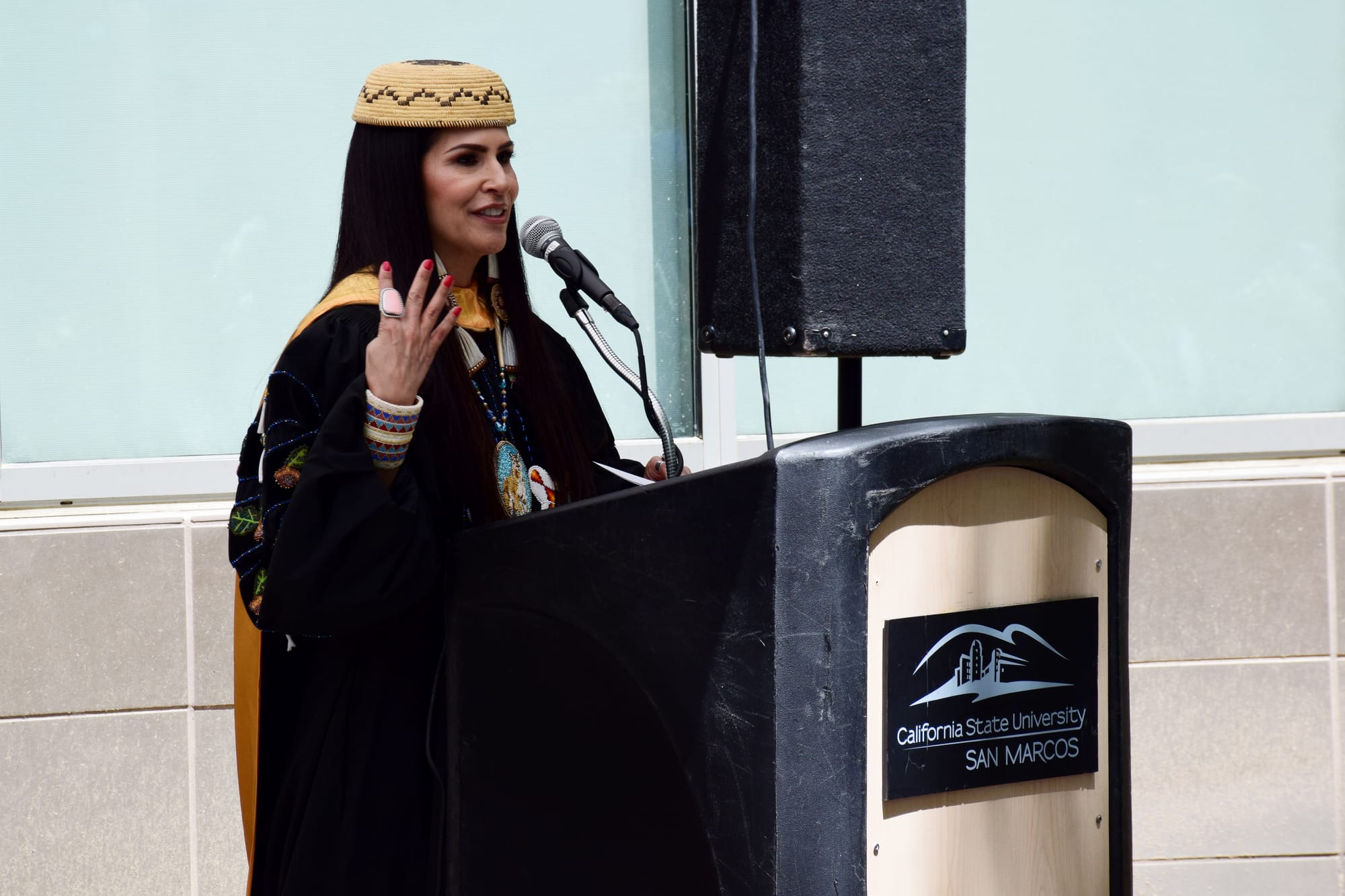 A woman with a woven hat and commencement robes speaks at a podium