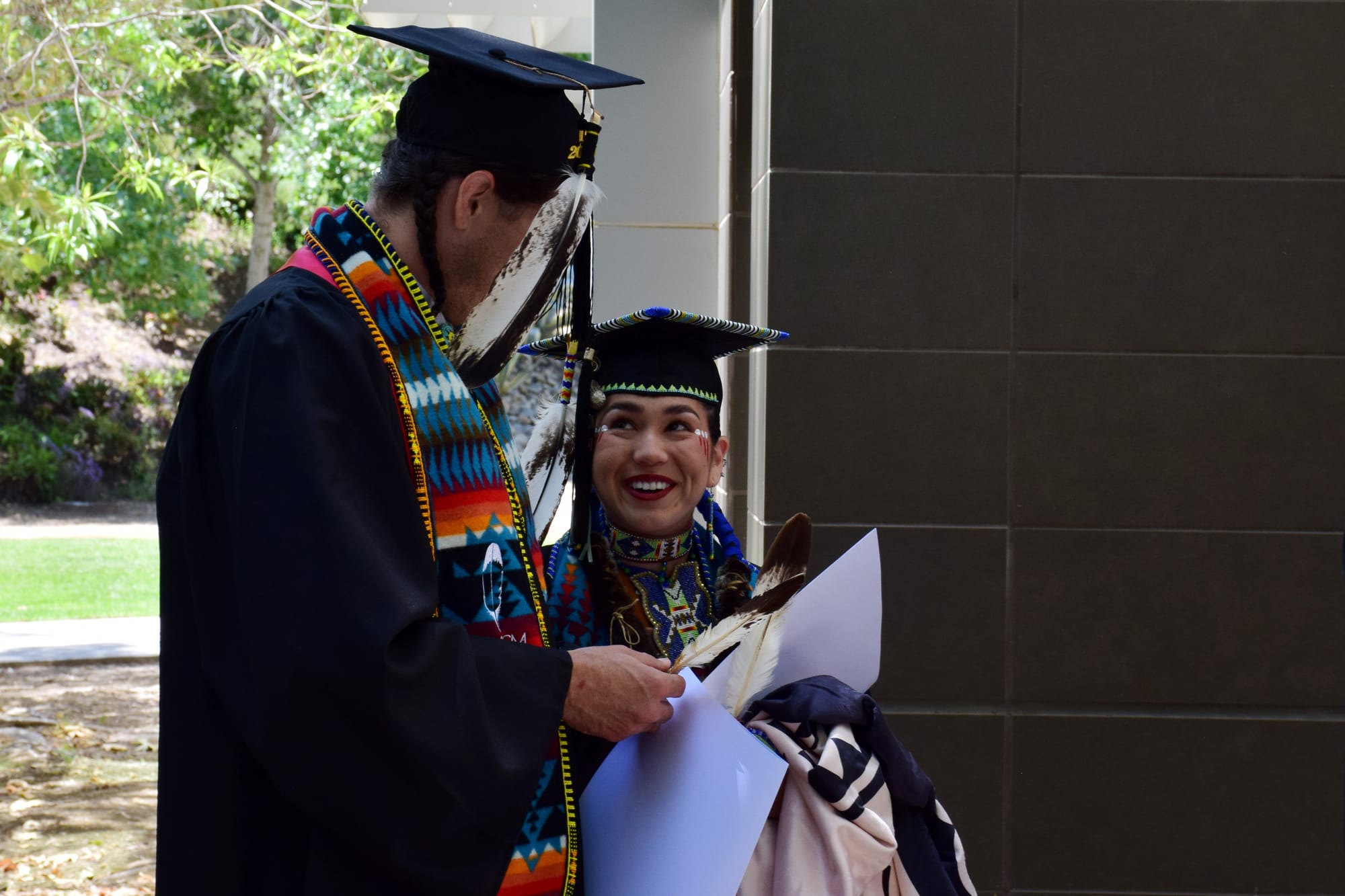 Two people in graduation regalia hold eagle feathers and certificates.