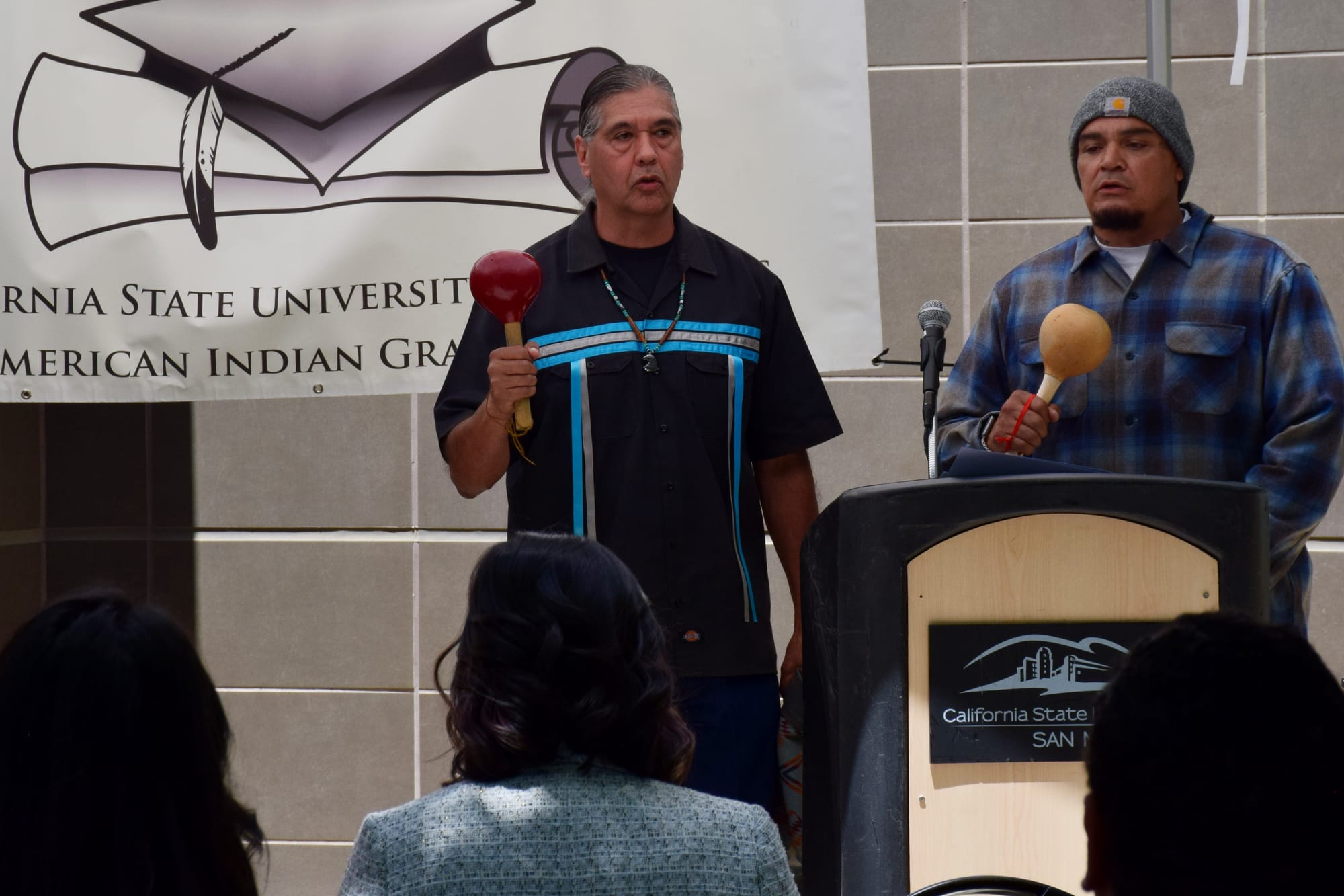 Two men stand in front od a crowd at an outdoor event singing and playing rattles