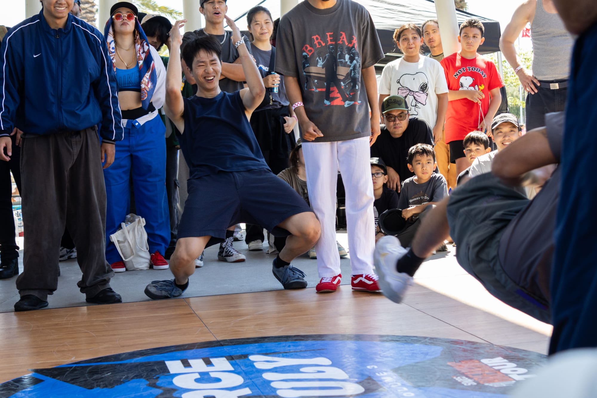 A dancer reacts to another dancer by dropping down to their knees in a breaking competition in a crowd outside in a park.