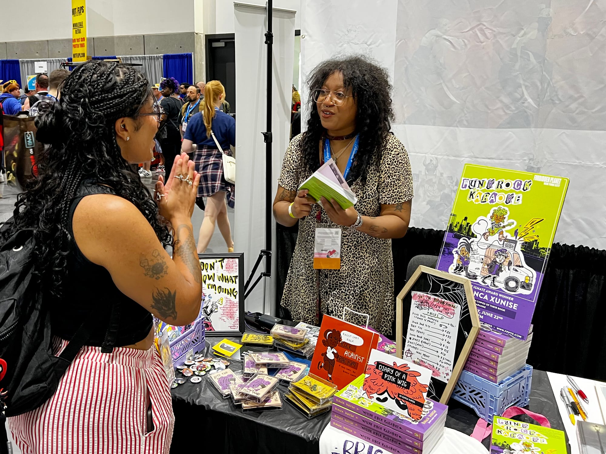 Two women talk at a comic book booth in a convention center 
