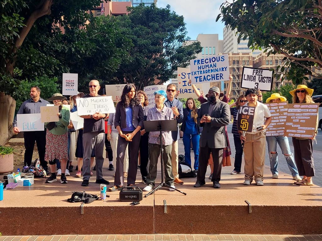 People stand with signs that read "precious human extraordinary teacher," "free Linda Vista 3" and "ICE out of SD" as someone speaks on a platform .