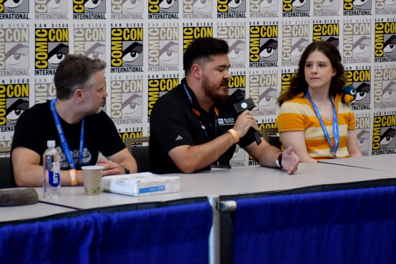 Three people sit at a panel with the comic-con logo on a backdrop behind them.