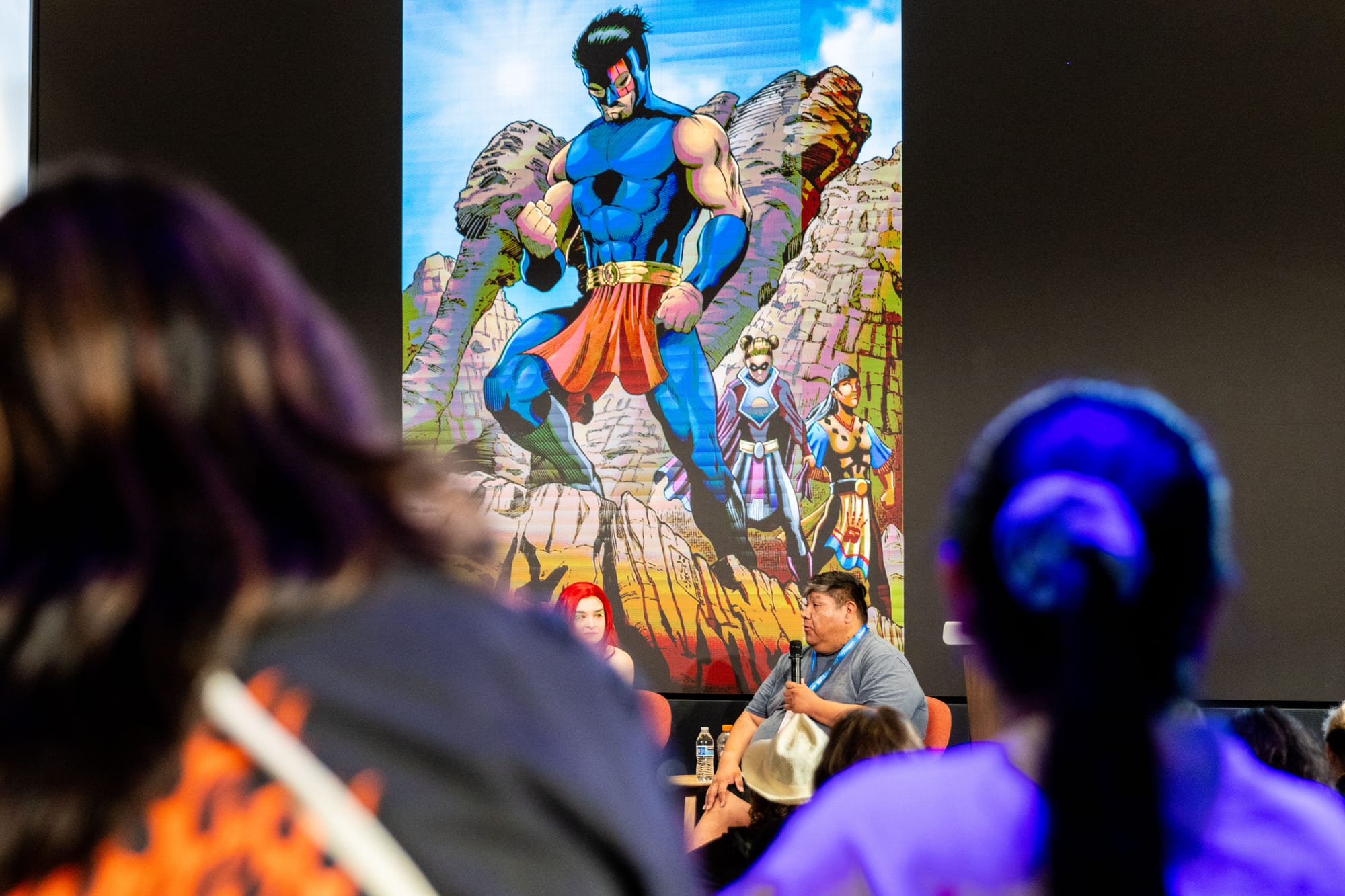 close up of the back of audience members' heads with a man speaking on stage and a comic book character on a screen behind him