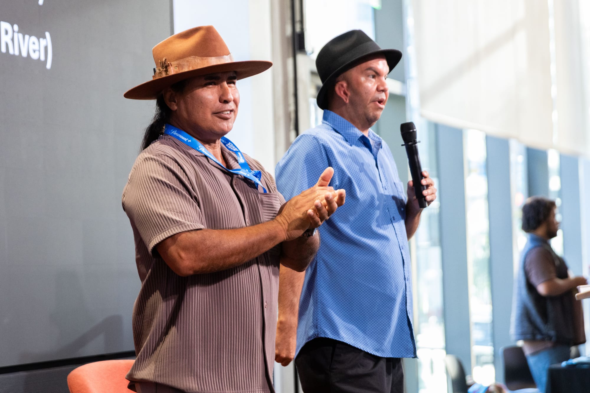 Two men wearing hats talk on a stage