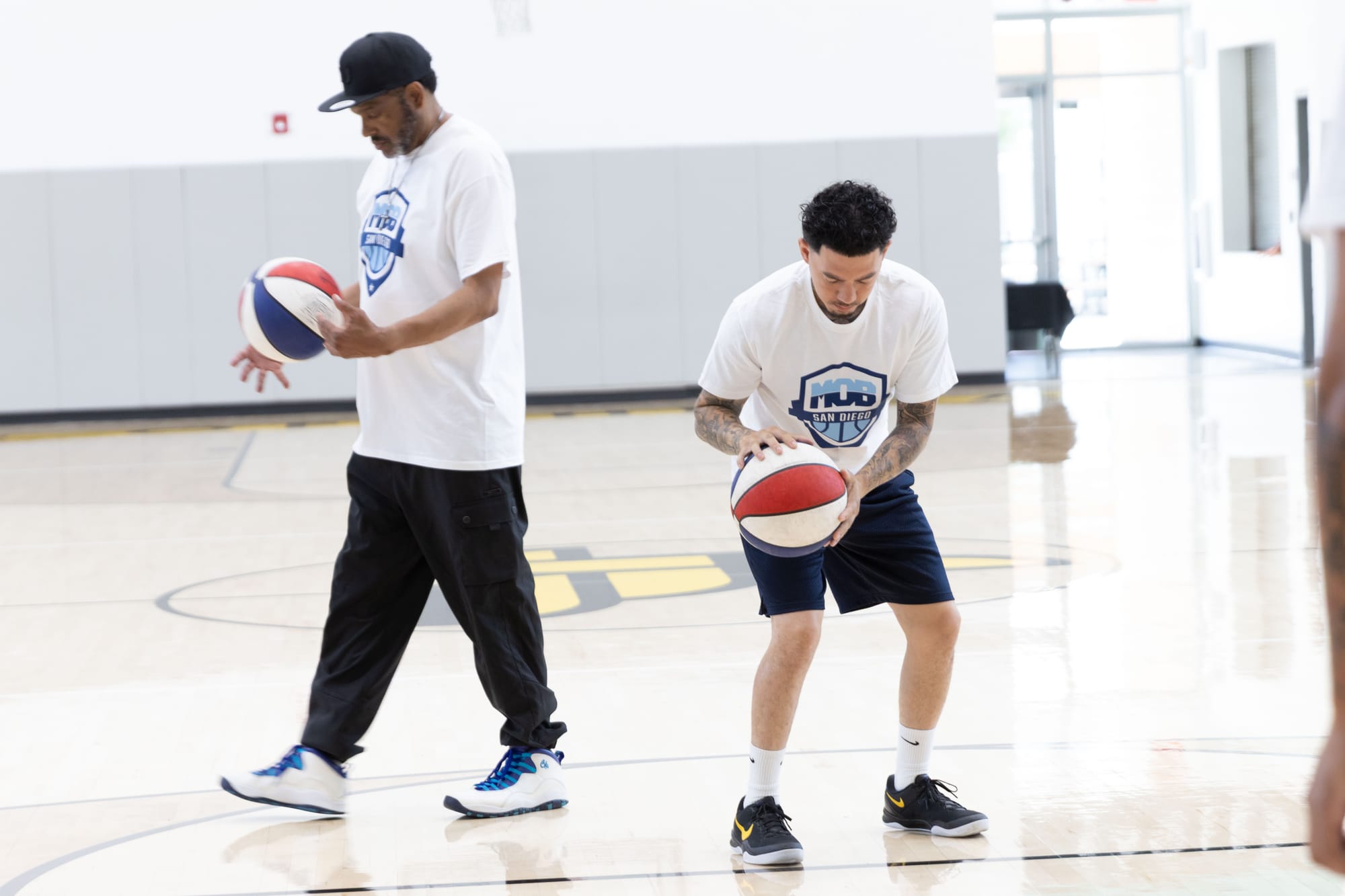 One person walks holding a basketball while another one dribbles a ball on a basketball court