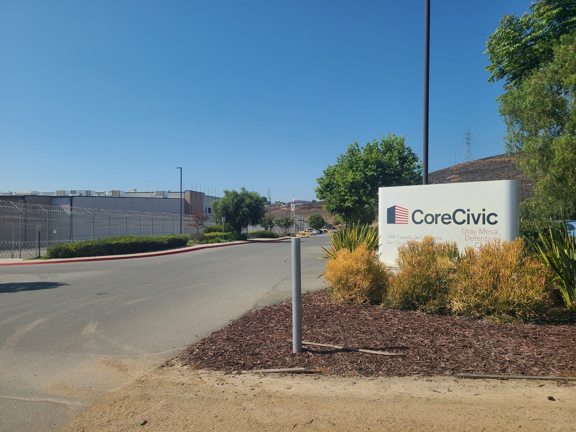 A prison-like building sits to the left of a parking lot and a sign that says CoreCivic Otay Mesa Detention Center