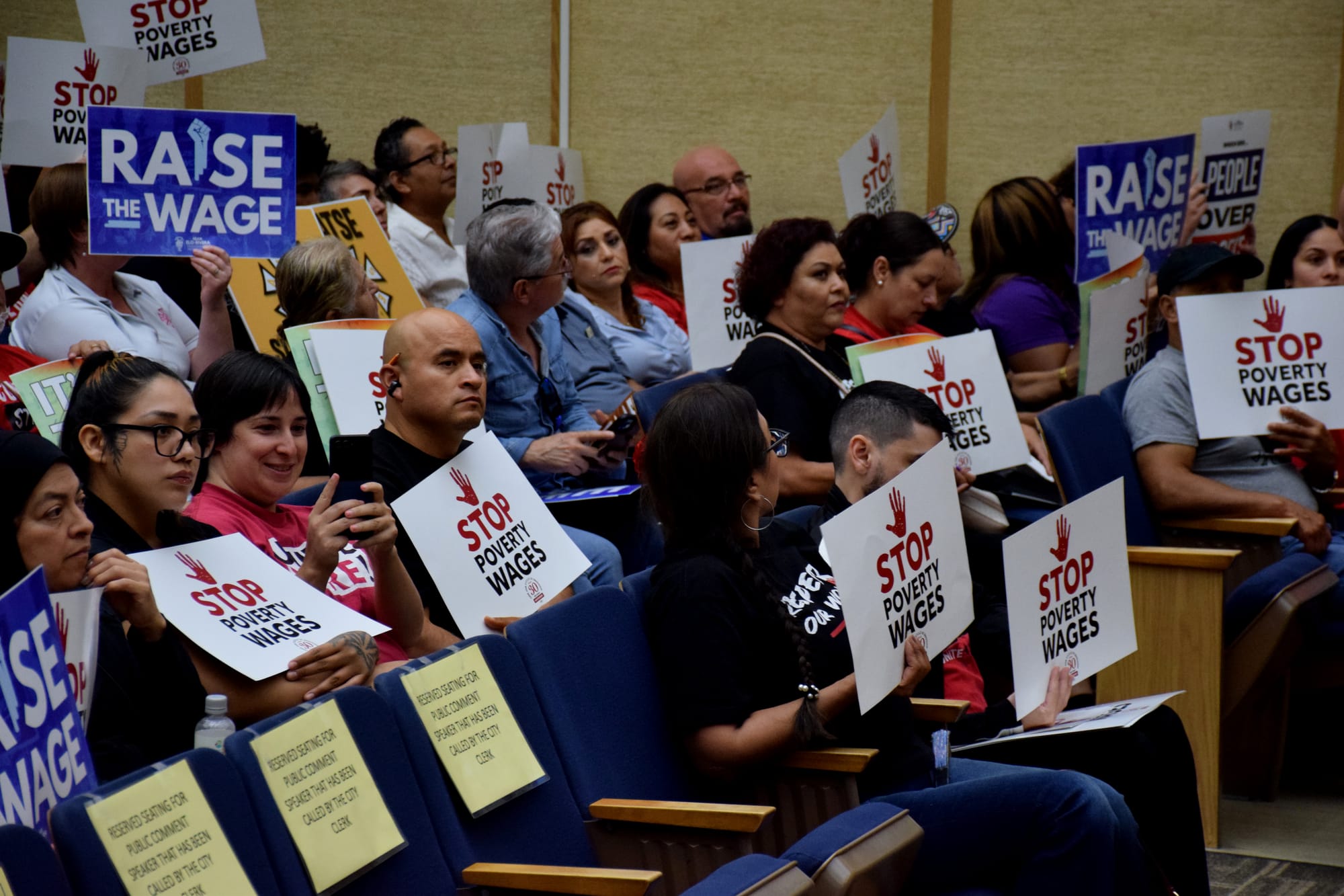 People watching Tuesday's San Diego City Council meeting hold signs that read "Stop Poverty Wages" and "Raise the Wage" 
