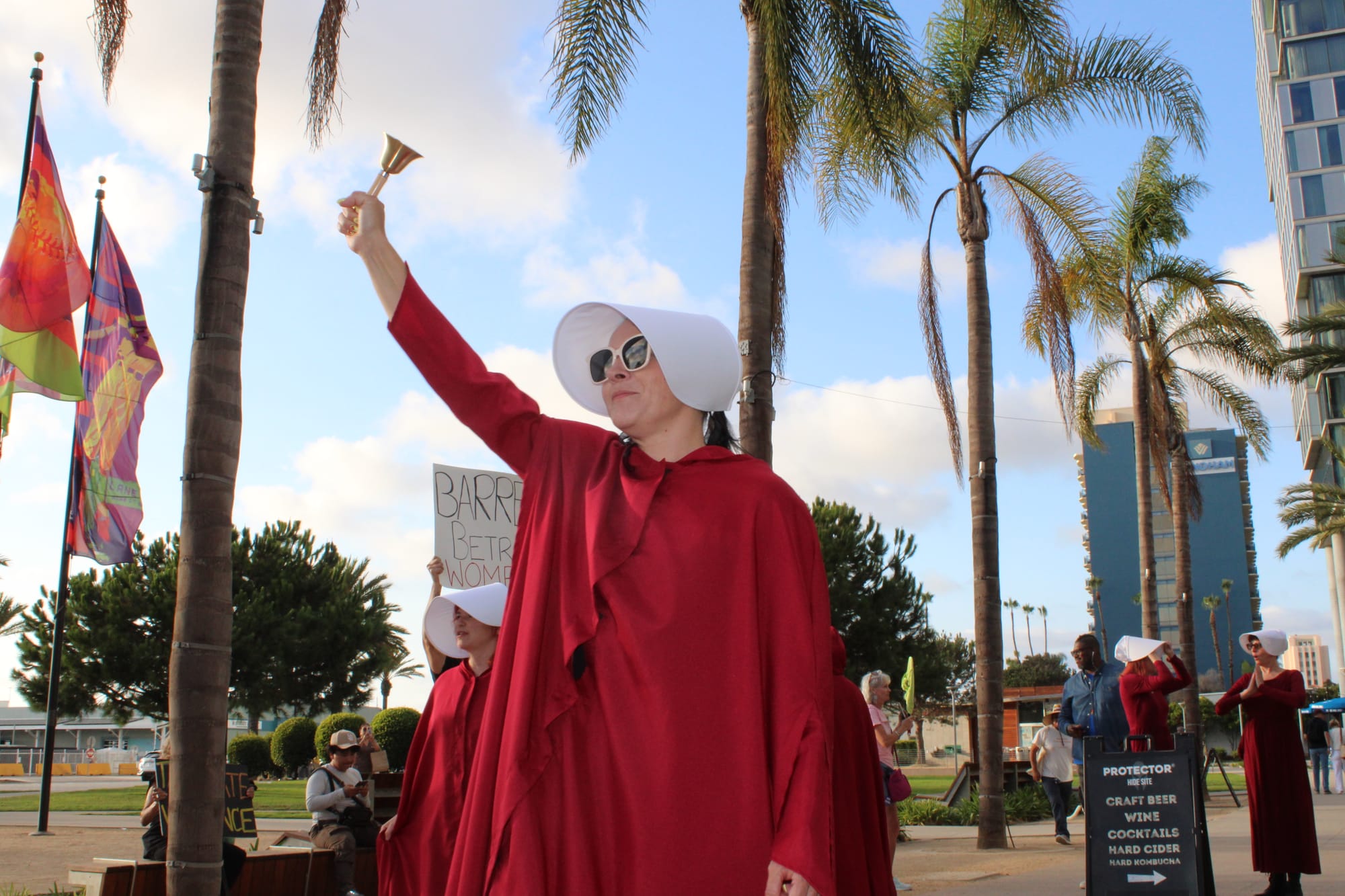 A woman in a red cape and white bonnet walks in a protest holding a bell in front of palm trees 