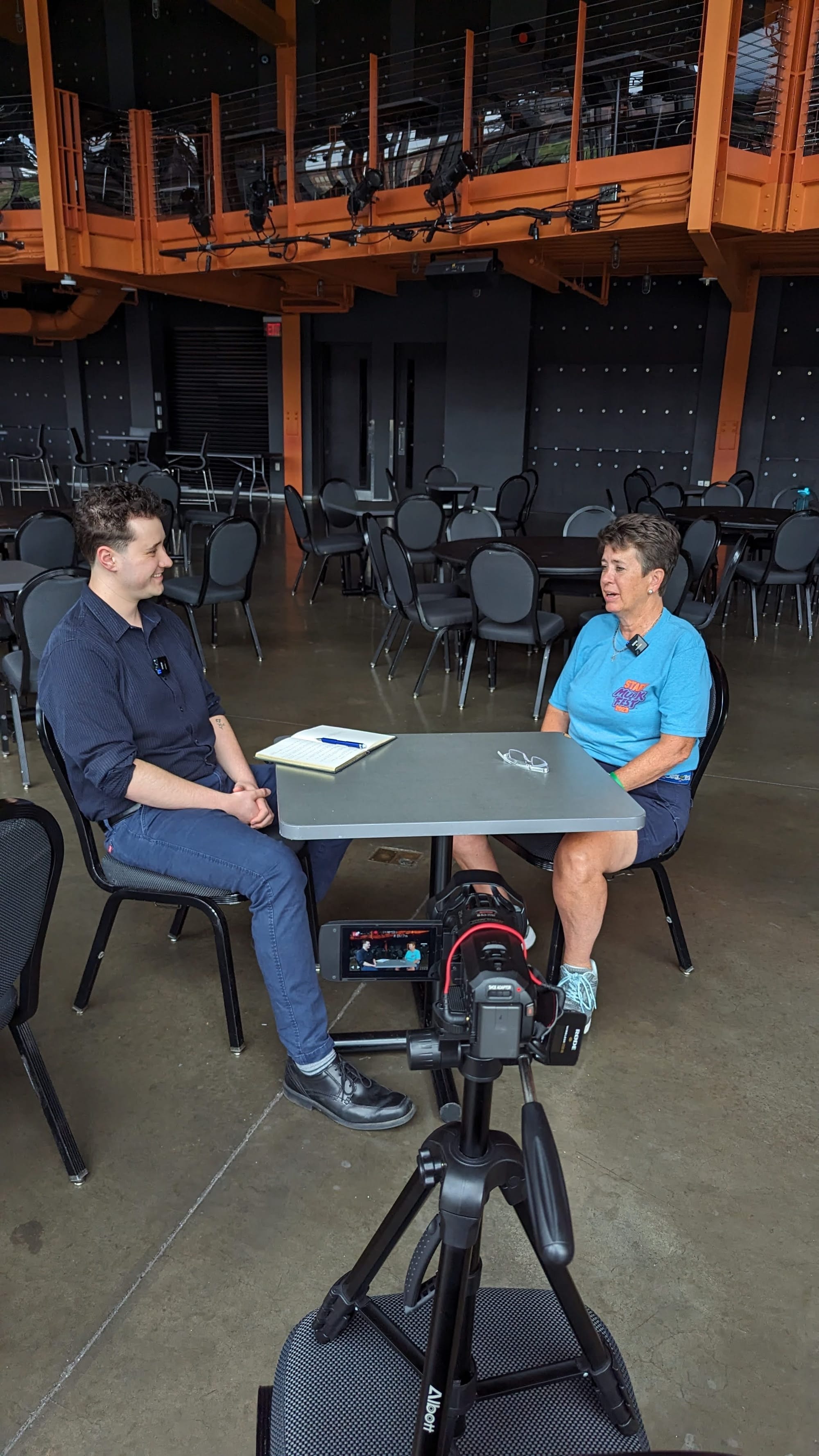 Two people sit a table talking during an interview with a camera in front of them