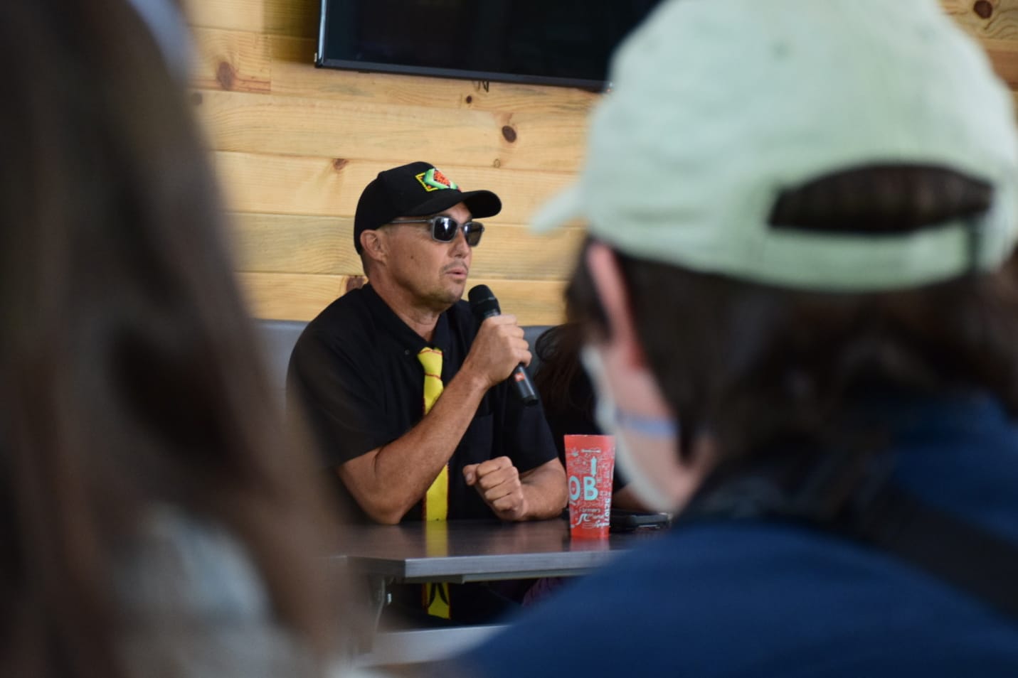 A man speaks into a microphone while sitting at a table. Two people are sitting with their backs to the camera in front of him