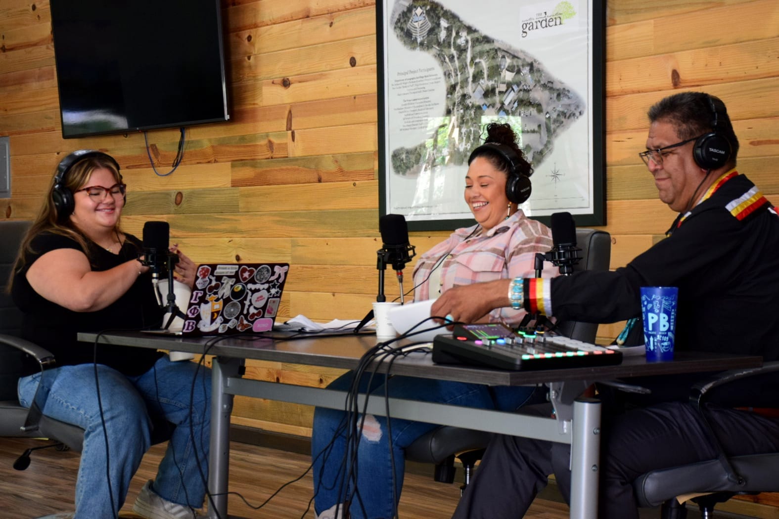 Three people sit in front of microphones with headphones on and a mixing board in front of them