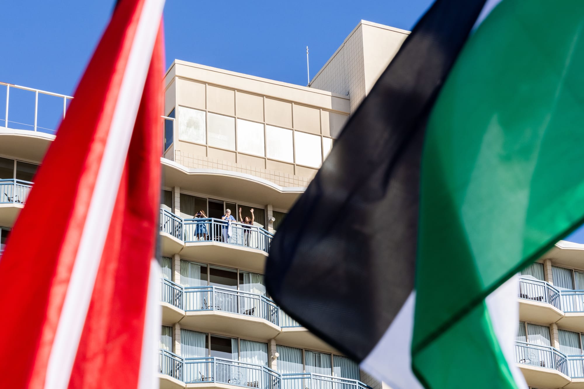 Closeup of palestinian flags with a building on the background and people waving from a balcony