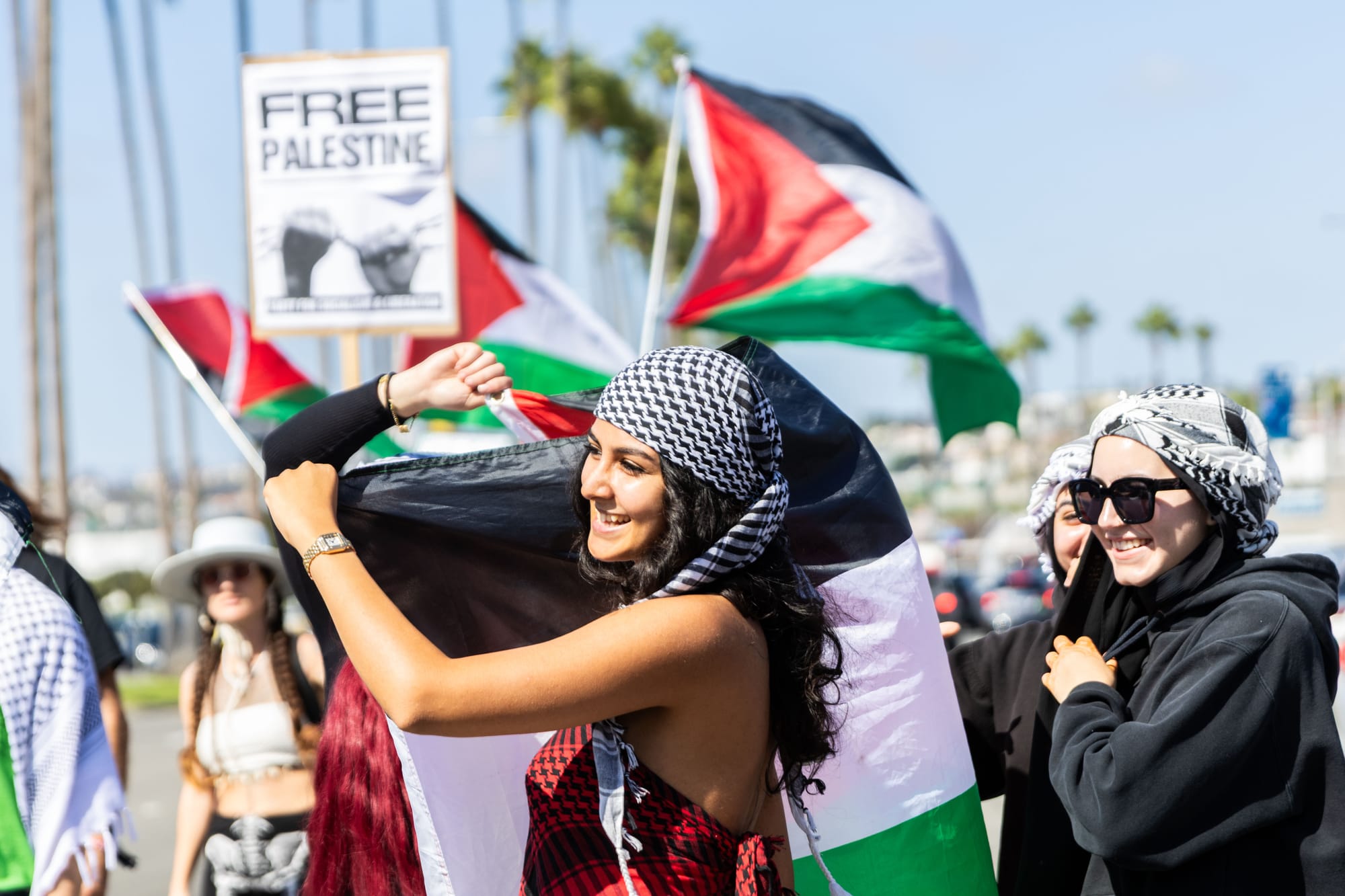 A group of people hold palestinian flags a a sign that reads "free palestine" during a march by the harbor with palm trees behind them