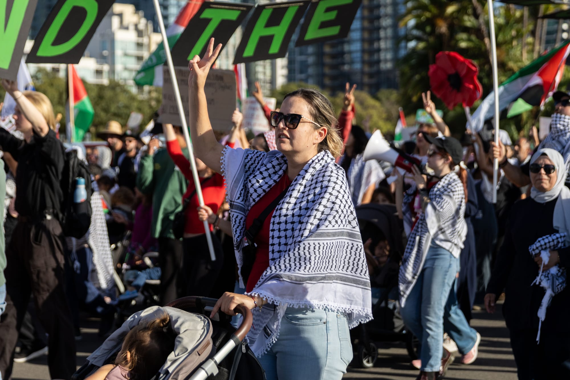 A group of people wearing keffiyehs and holding palestinian flags hold up peace sides with their fingers while walking in a march