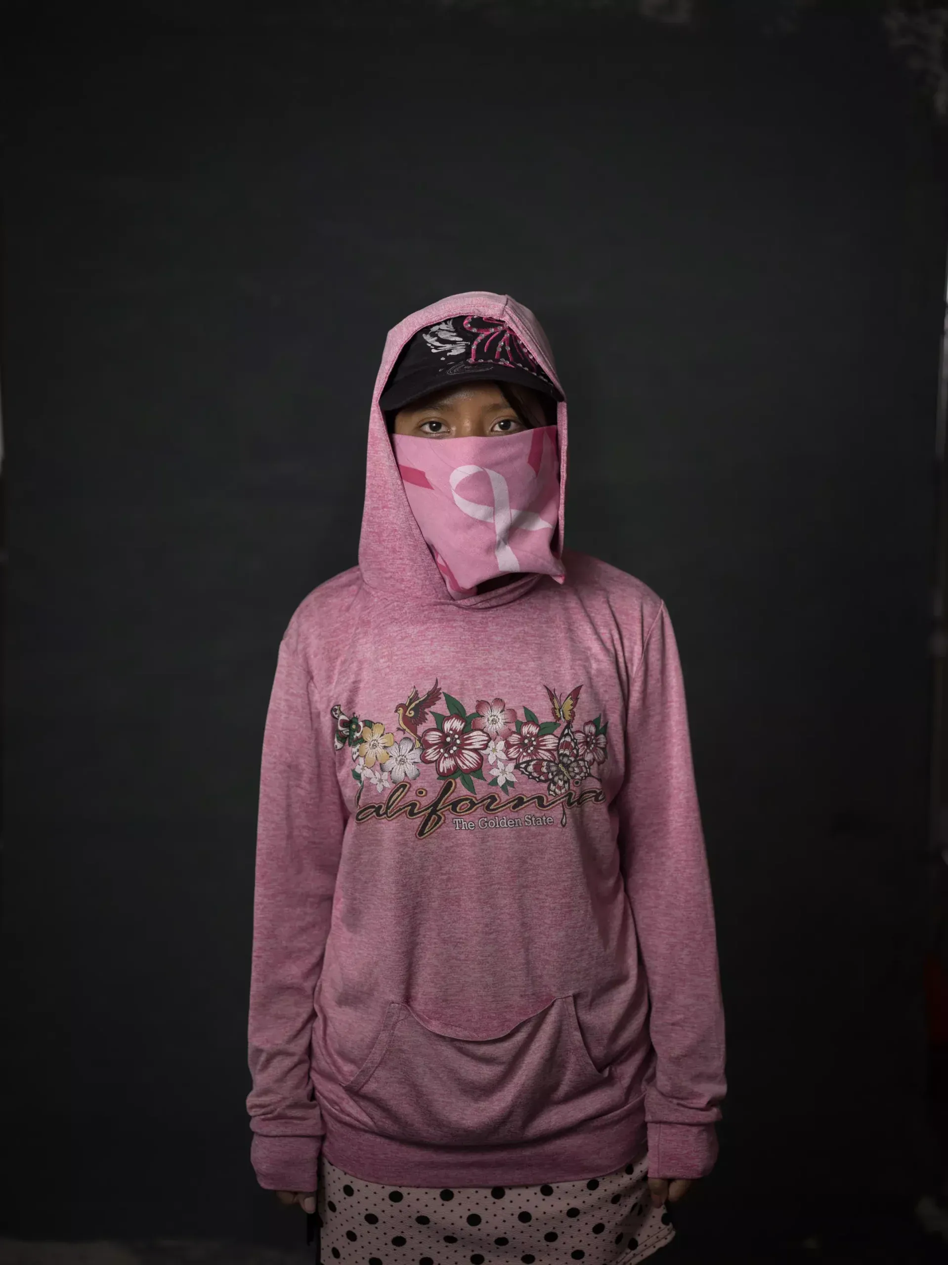 A girl stands in a pink shirt with a hood and a pink bandana across her face
