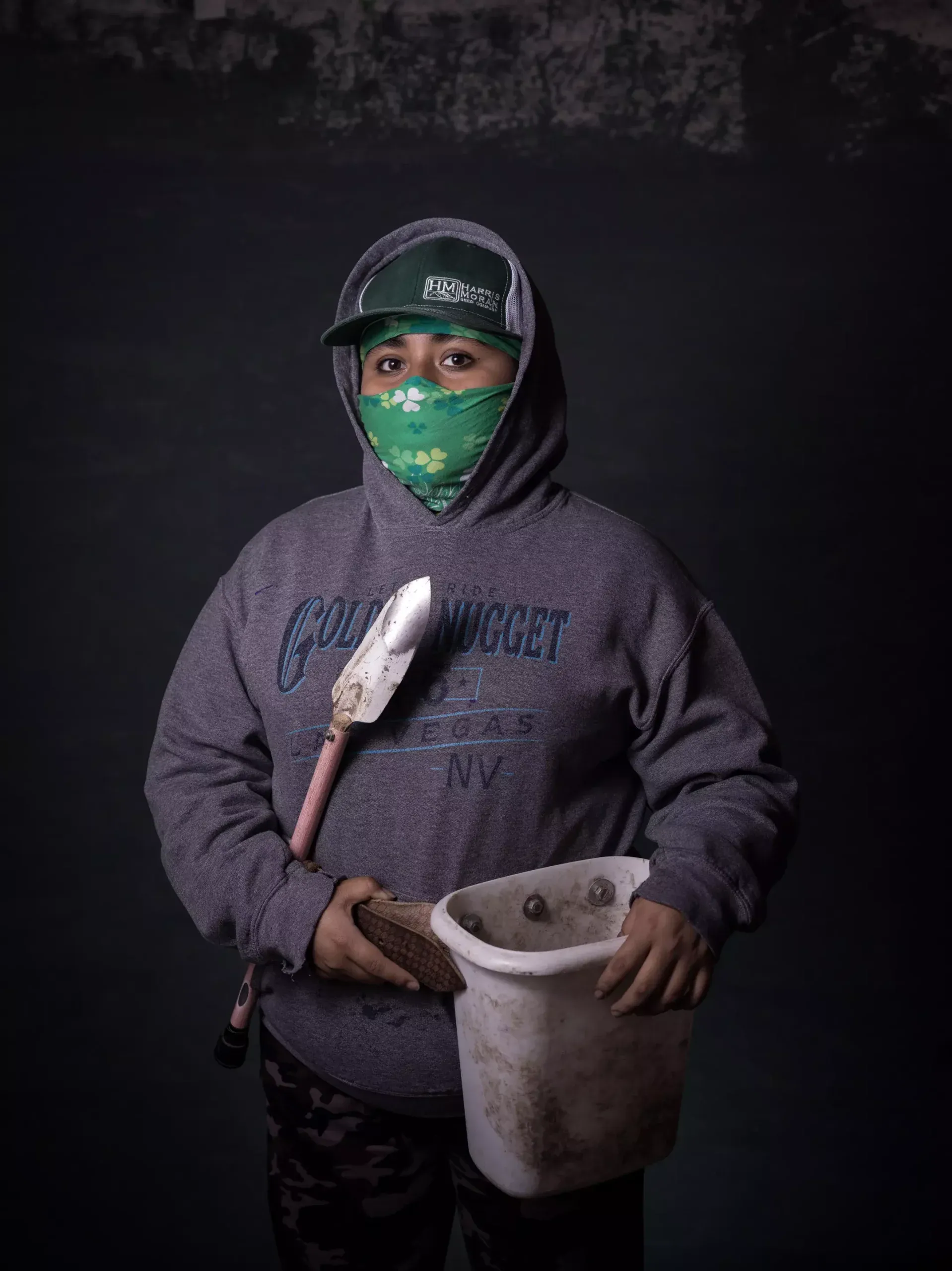 A girl in a hoodie and bandana holds a shovel and bucket