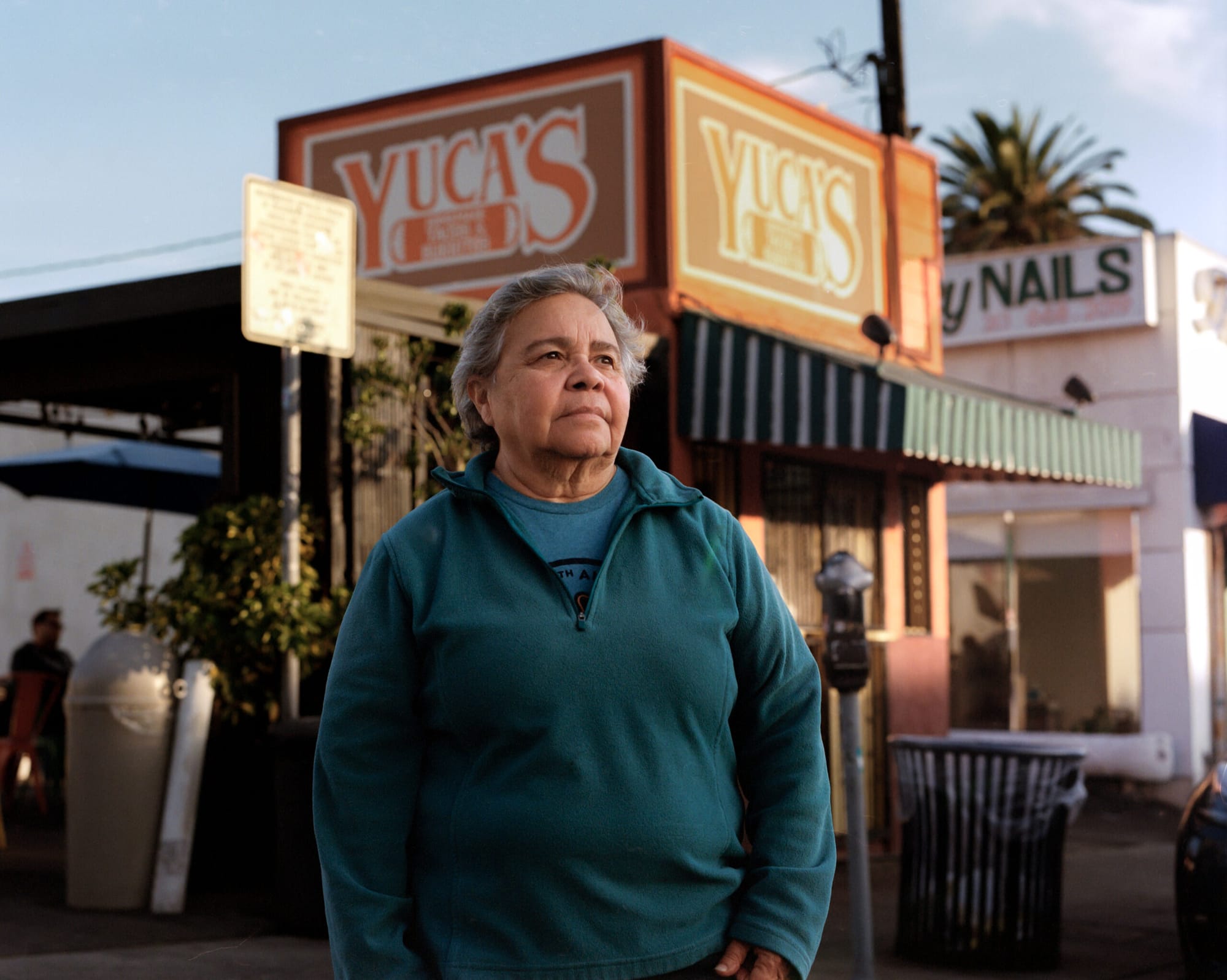 A woman in a green sweater stands in front of a restaurant with a sign that says yuca's