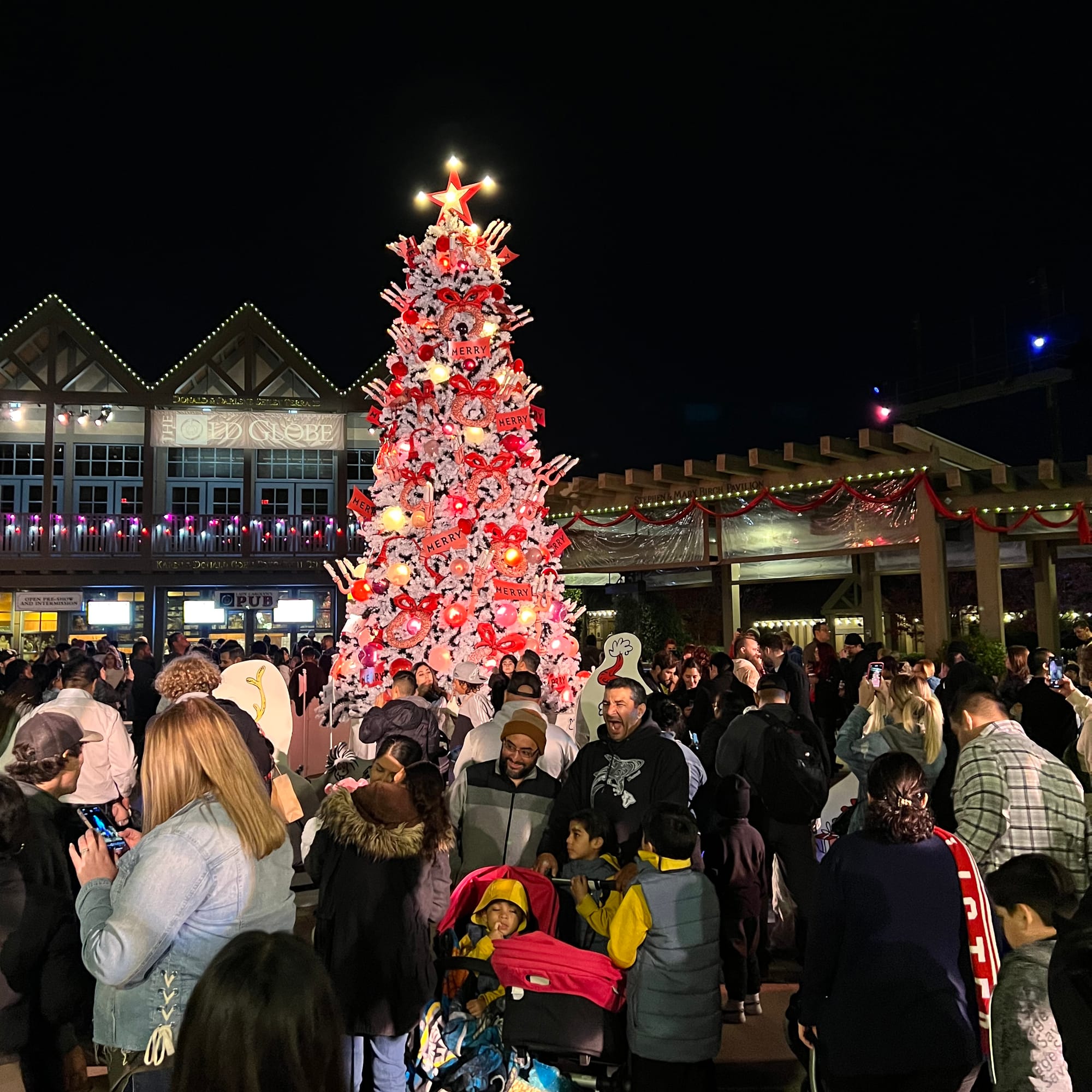 Picture of a vibrant pink, red and white Christmas tree with ornate decorations and a crowd of people standing around it outside.
